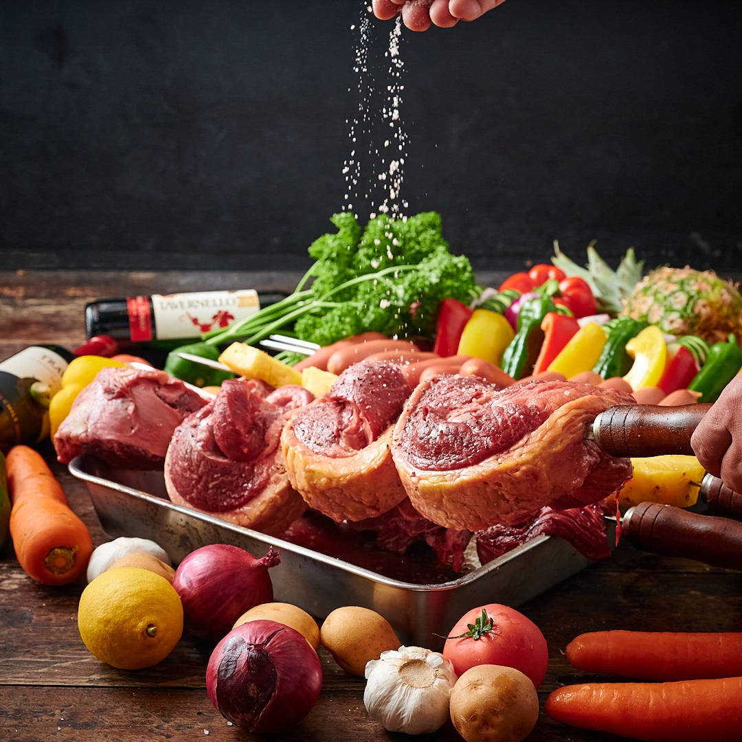 香料調味的炭烤肉，即將上烤架 壁紙 — 一隻手在質樸的木桌上，將鹽撒在被色彩繽紛的新鮮蔬菜包圍的生牛肉烤肉上。