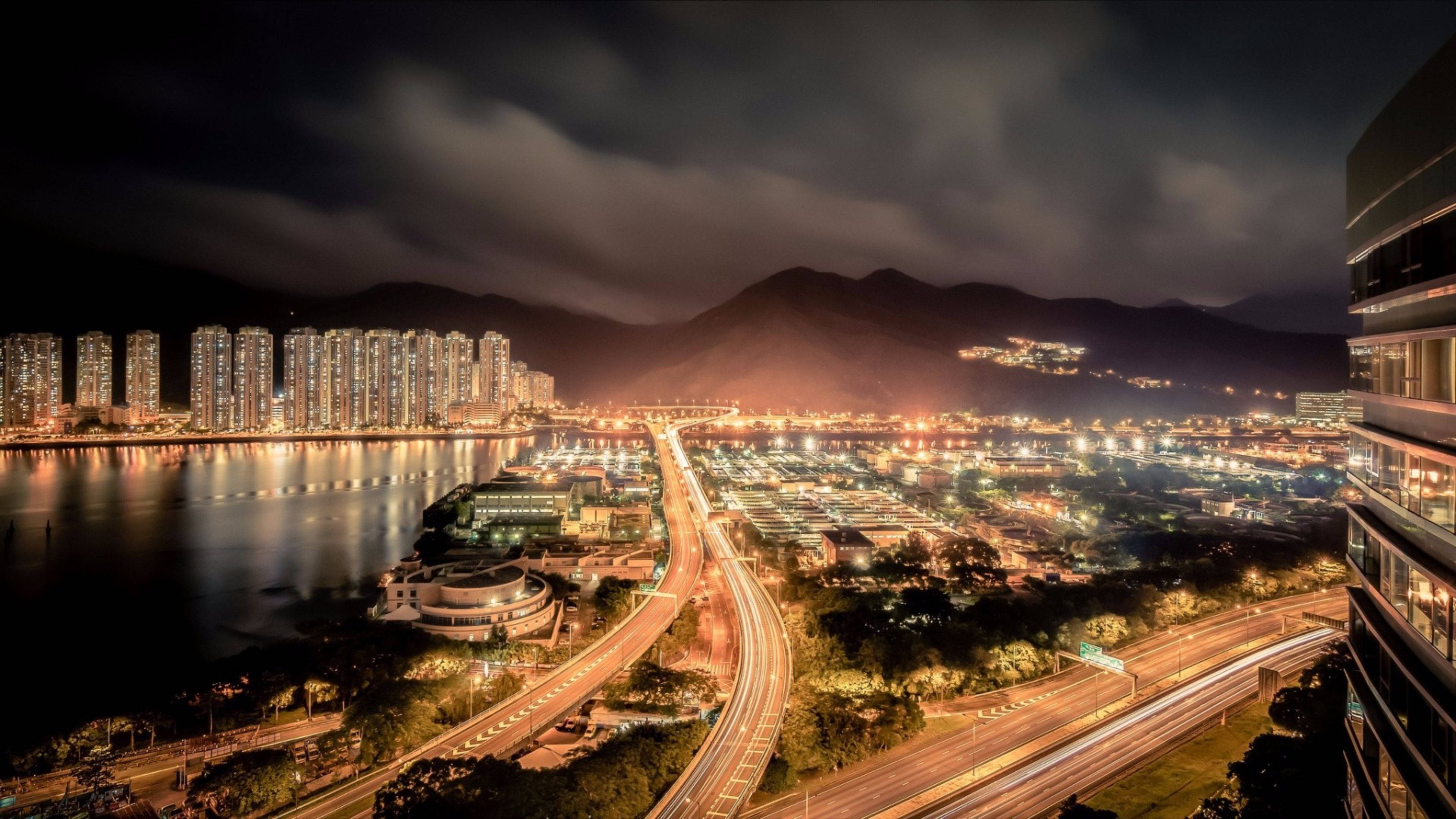 香港城市燈光熱門圖 壁紙 — 一個充滿活力的夜景城市景觀，有發光的摩天大樓、照明的高速公路和遠處的山脈，天空多雲。