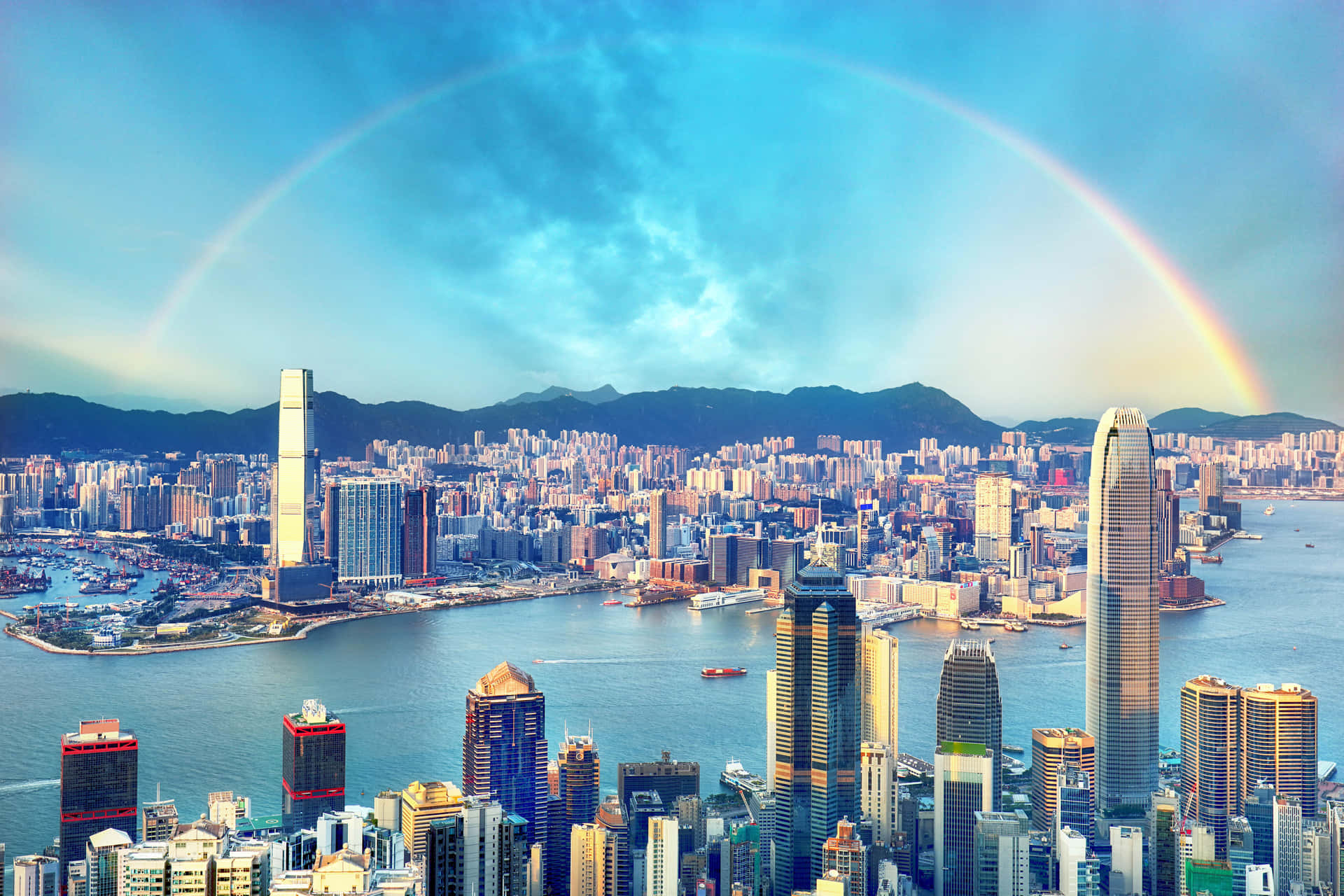 香港城市璀璨夜景 壁紙 — 一道鮮豔的彩虹橫跨香港密集的天際線和港口，背景有山巒。