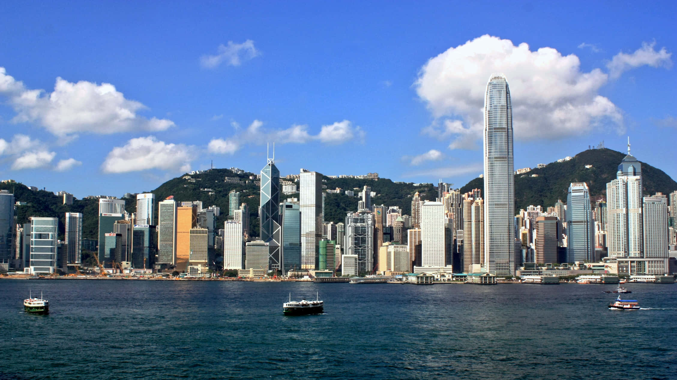 香港夜間天際線盛景 壁紙 — 香港天際線的全景視圖，高聳的摩天大樓和港口的船隻在明亮的藍天白雲下。