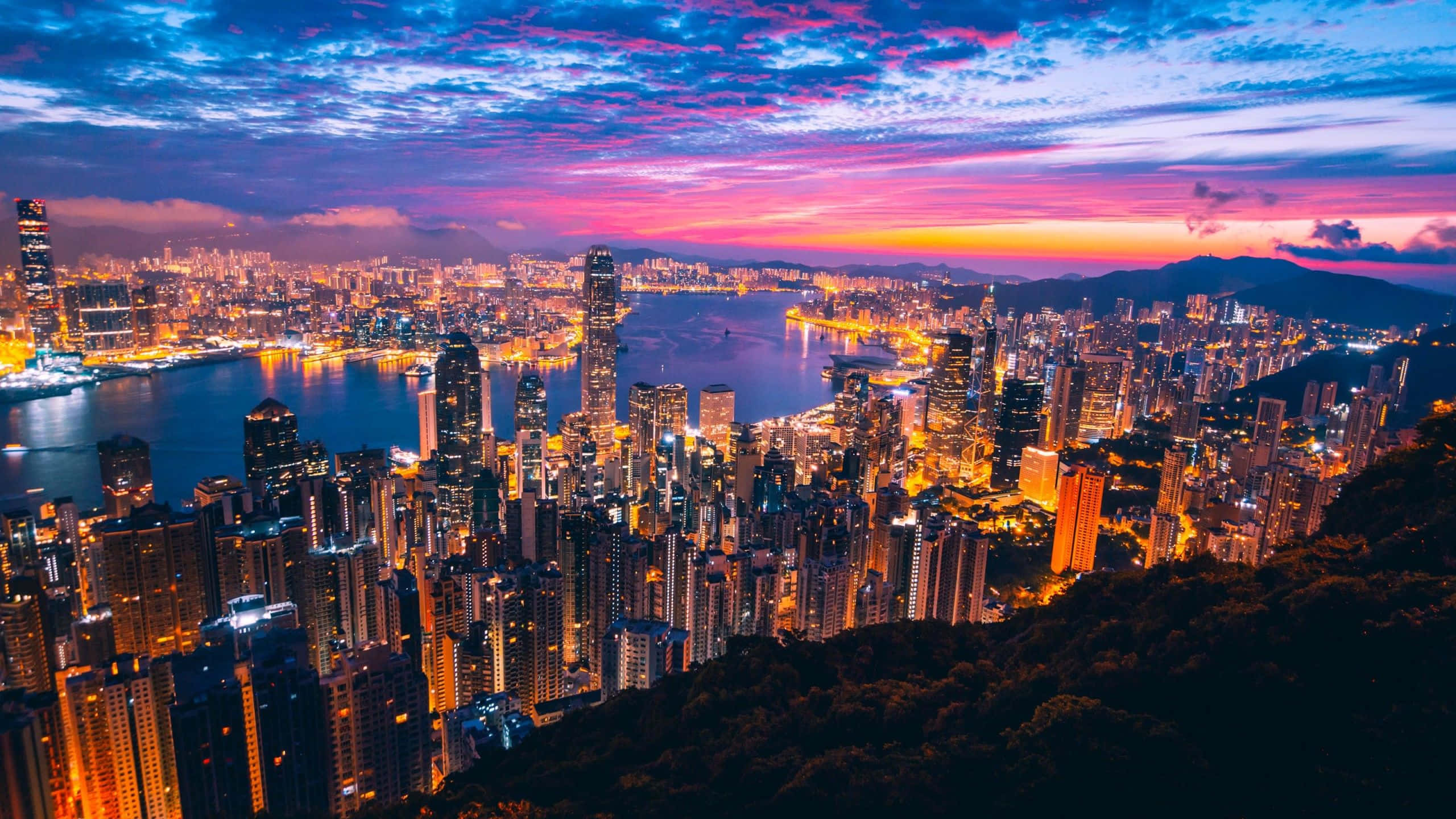 View 桌布 壁紙 — 香港天際線在暮色中令人驚嘆的景色，閃爍的摩天大樓在充滿活力的紫色和橙色天空下倒映在港口中。