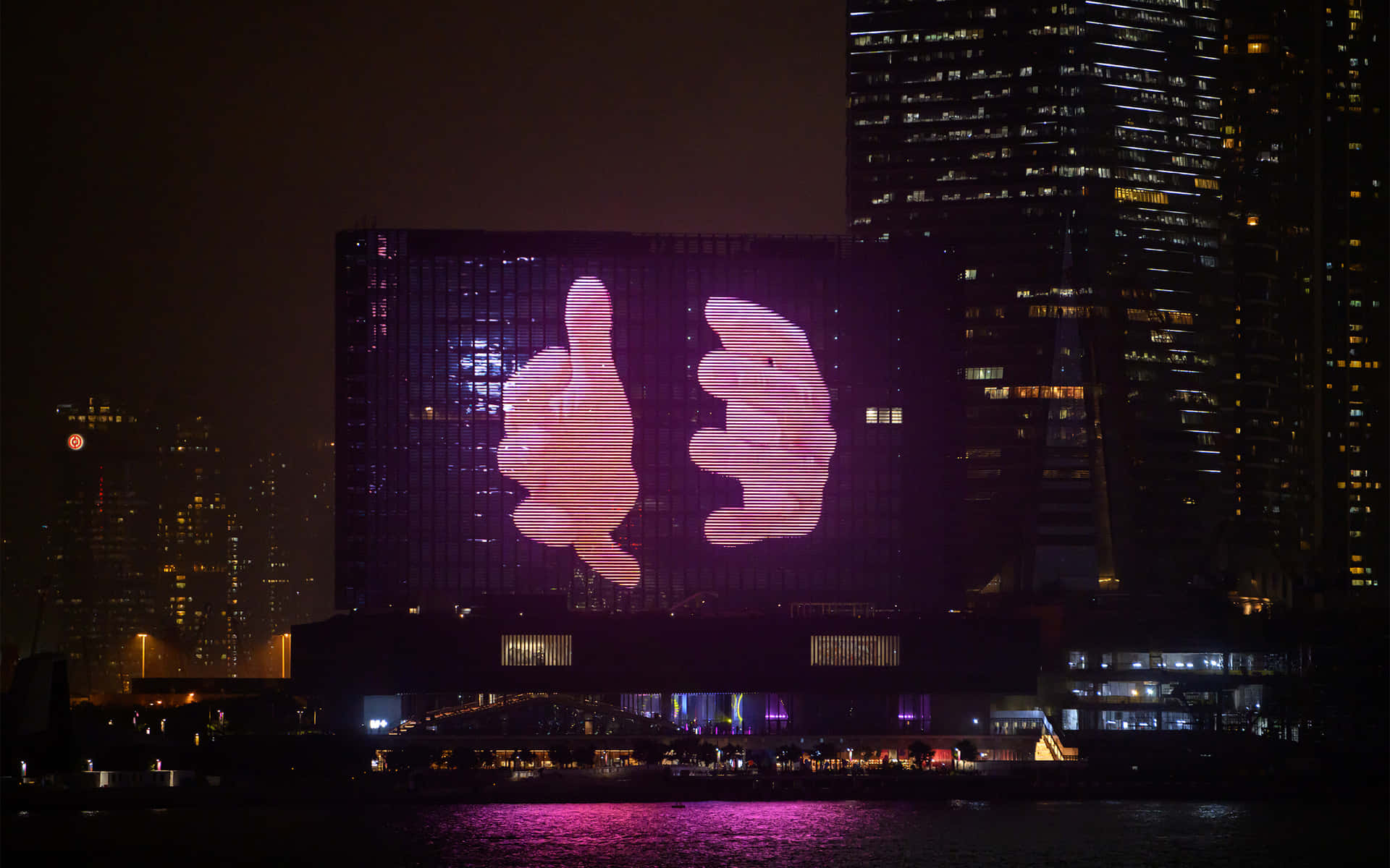 香港天際線黃昏美景 壁紙 — 夜晚，一座巨大建築物的外牆在 illuminated 摩天大樓和黑暗水面倒影的背景下，顯示出一個發光的粉紅色豎起大拇指和放下大拇指的手勢。