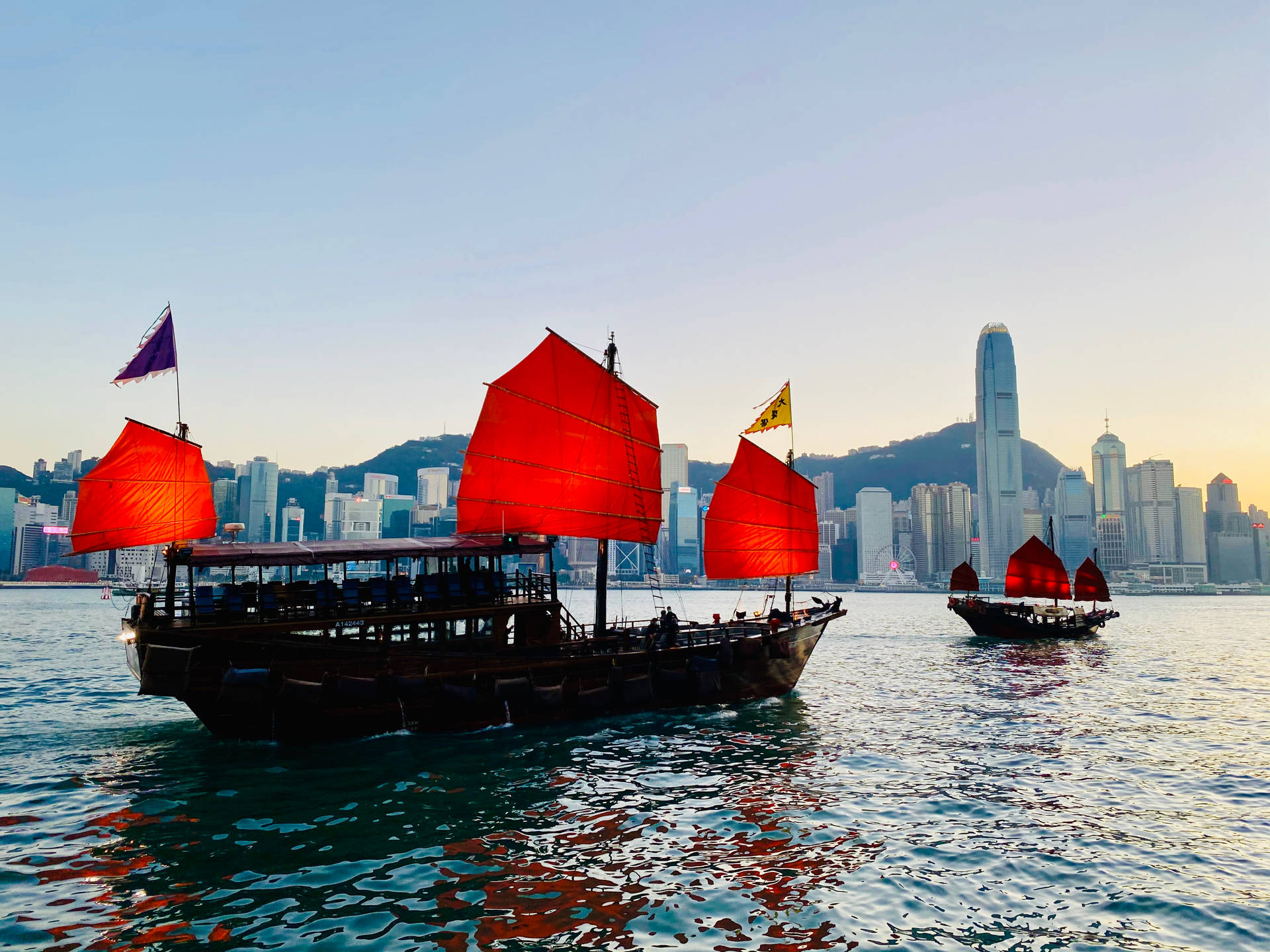香港鴨脷洲遊船 壁紙 — 傳統紅帆帆船在平靜的水面上滑行，香港標誌性的天際線在金色夕陽中閃耀。