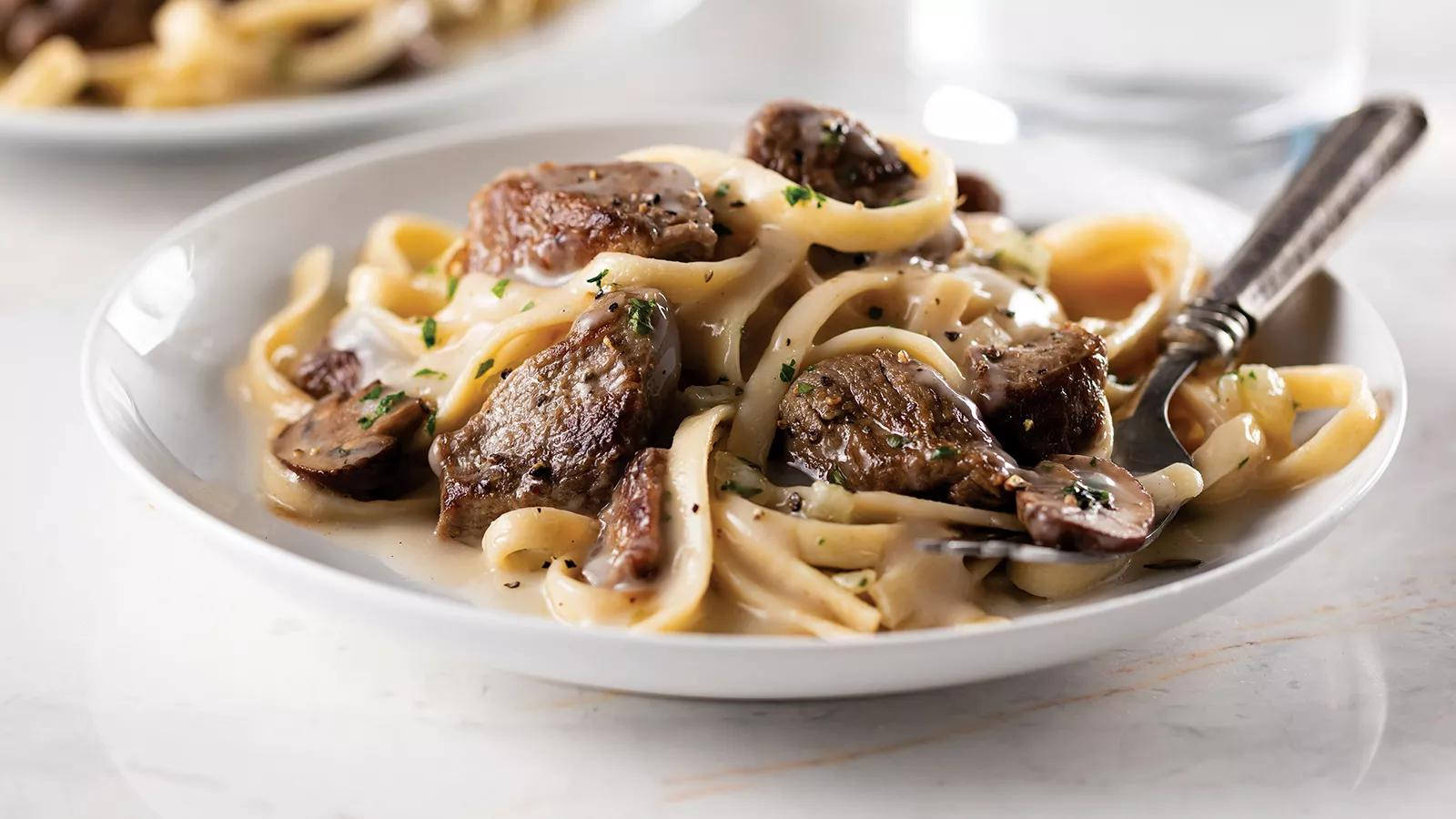 香濃牛肉斯特羅甘諾夫配 Linguine 意大利麵 壁紙 — 一個白色碗的特寫，裡面裝滿了奶油牛肉斯特羅加諾夫，放在寬麵條上，並用新鮮歐芹裝飾。