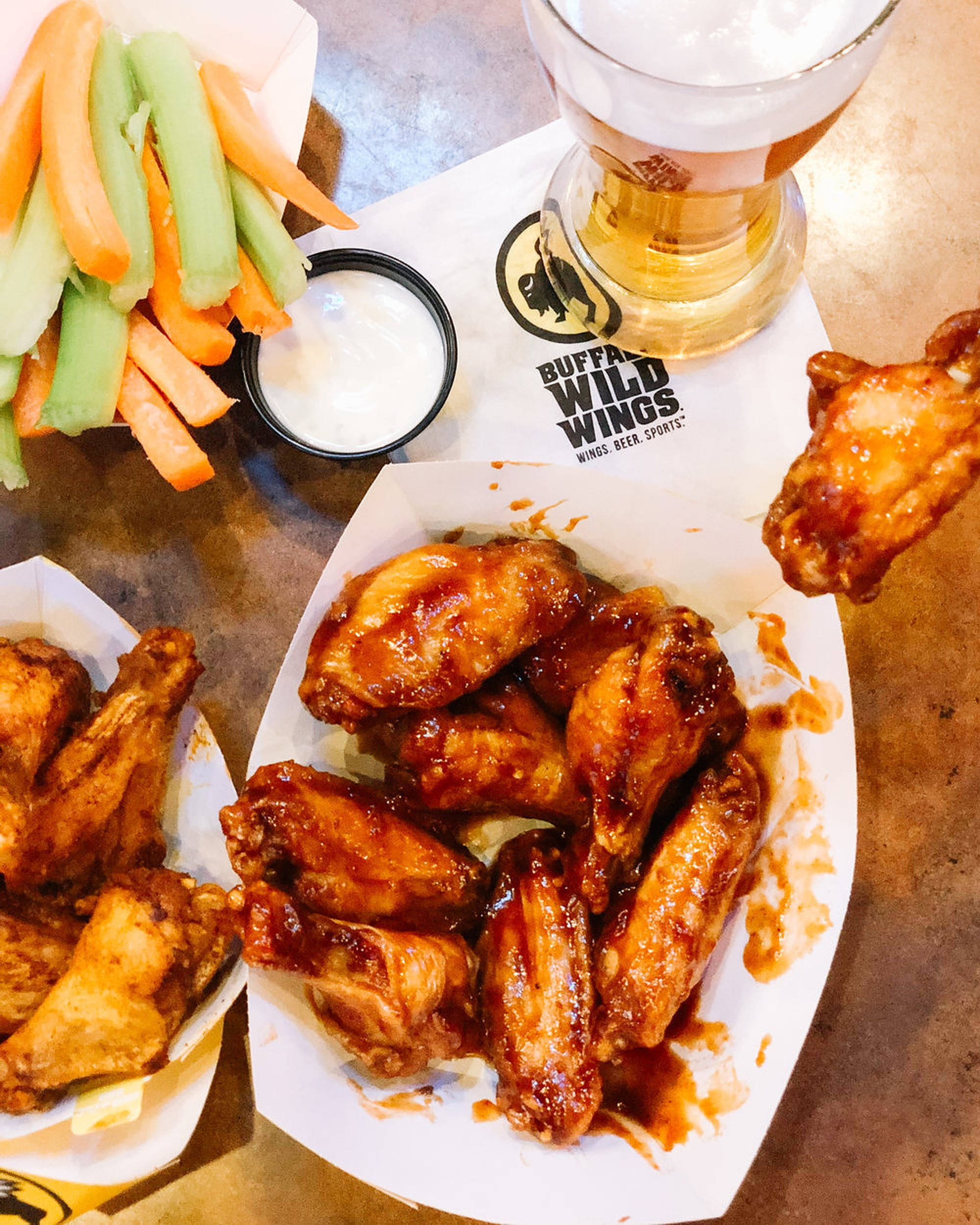 香辣 Buffalo Wild Wings 壁紙 — 閃亮的水牛城雞翼配西芹和紅蘿蔔條，旁邊放著一杯泡沫啤酒，放在有品牌標誌的紙托盤上。