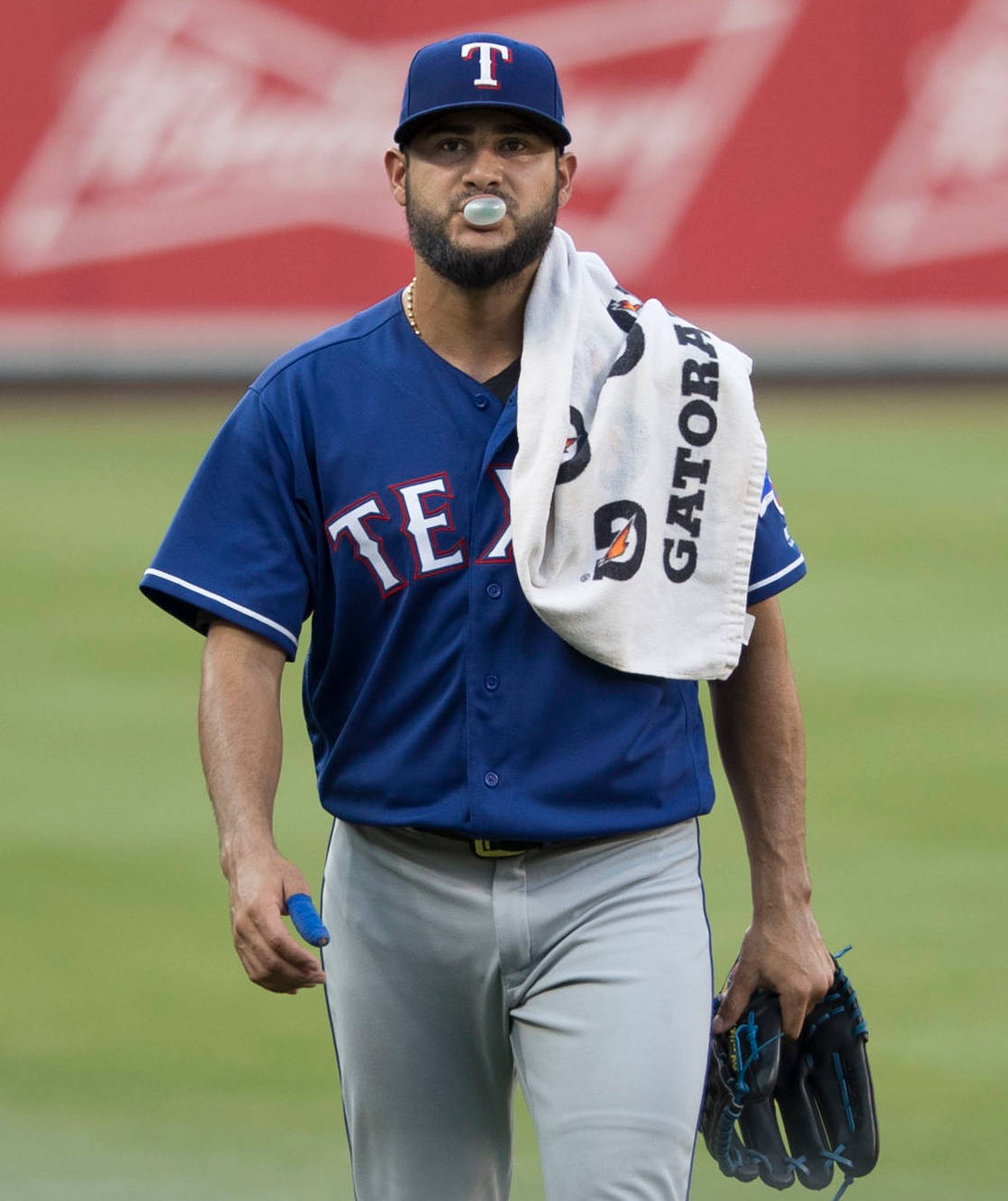 身穿德州遊騎兵隊球衣的棒球運動員在球場上行走，嘴裡吹著泡泡糖，肩上披著毛巾。