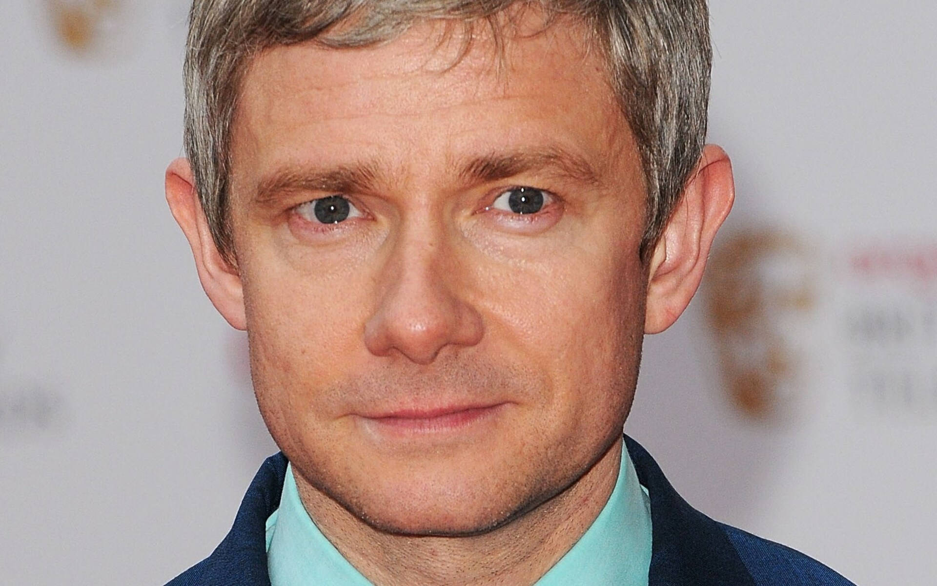 馬丁·弗里曼近照高清 壁紙 — Martin Freeman 的頭像特寫，灰白頭髮，表情平靜，身穿藍色西裝和青綠色襯衫，背景是模糊的活動場景。