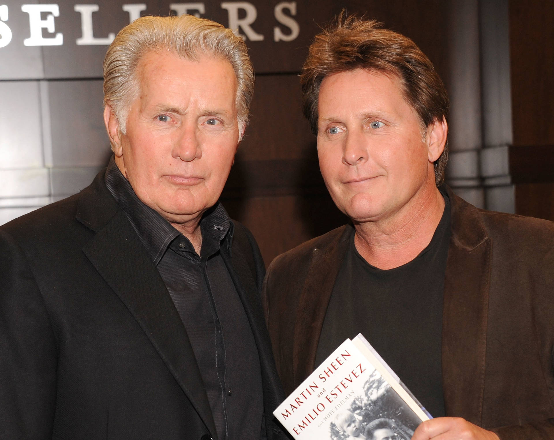 兩個男人在活動中擺姿勢，其中一人手持一本名為《Martin Sheen and Emilio Estevez》的書。