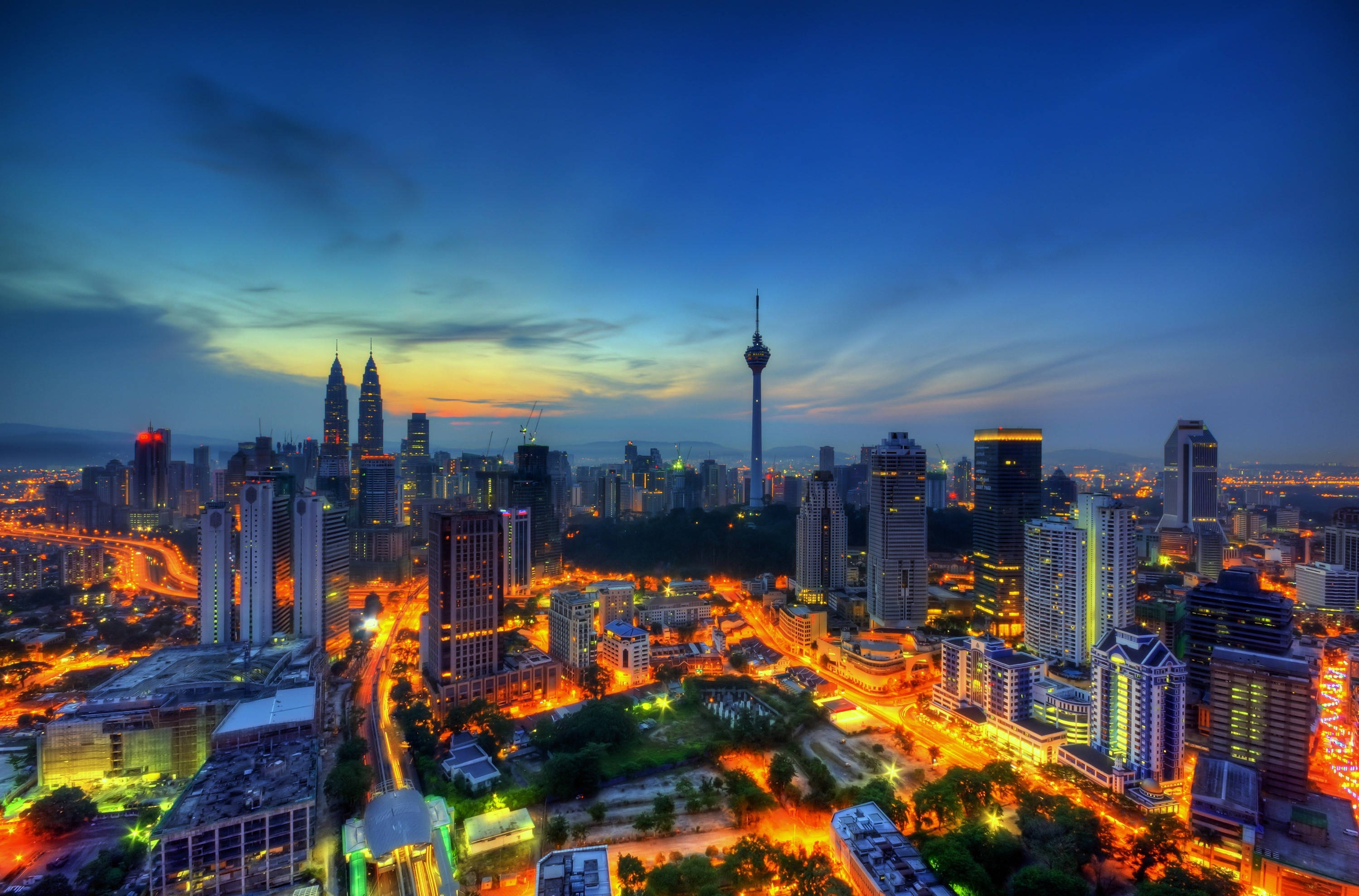 馬來西亞吉隆坡塔 壁紙 — 從高空俯瞰吉隆坡在黃昏時分的城市景觀，摩天大樓燈火通明，街道閃爍，深藍色的天空下顯得格外迷人。