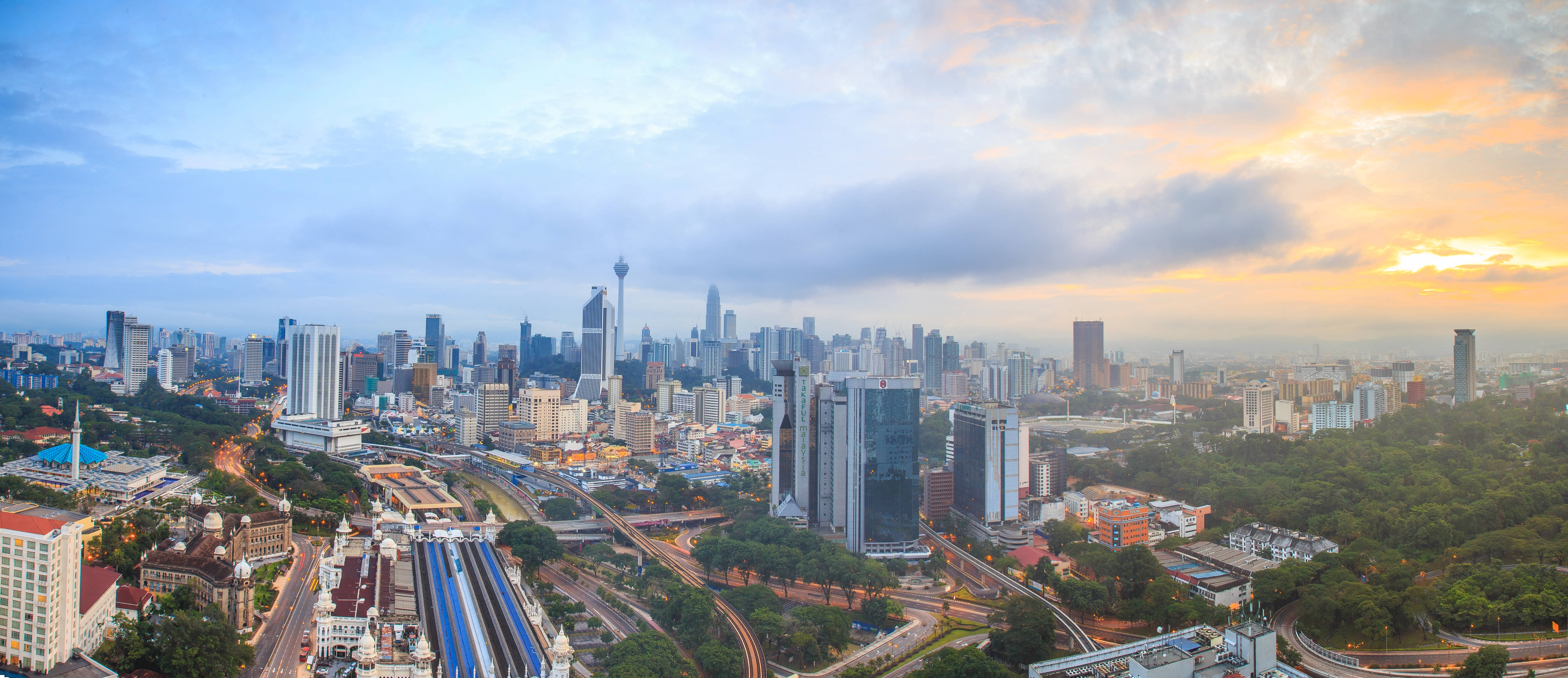 馬來西亞城市航拍 壁紙 — 現代城市天際線的 panoramic 景觀，夕陽西下，金色雲彩和高聳的摩天大樓。