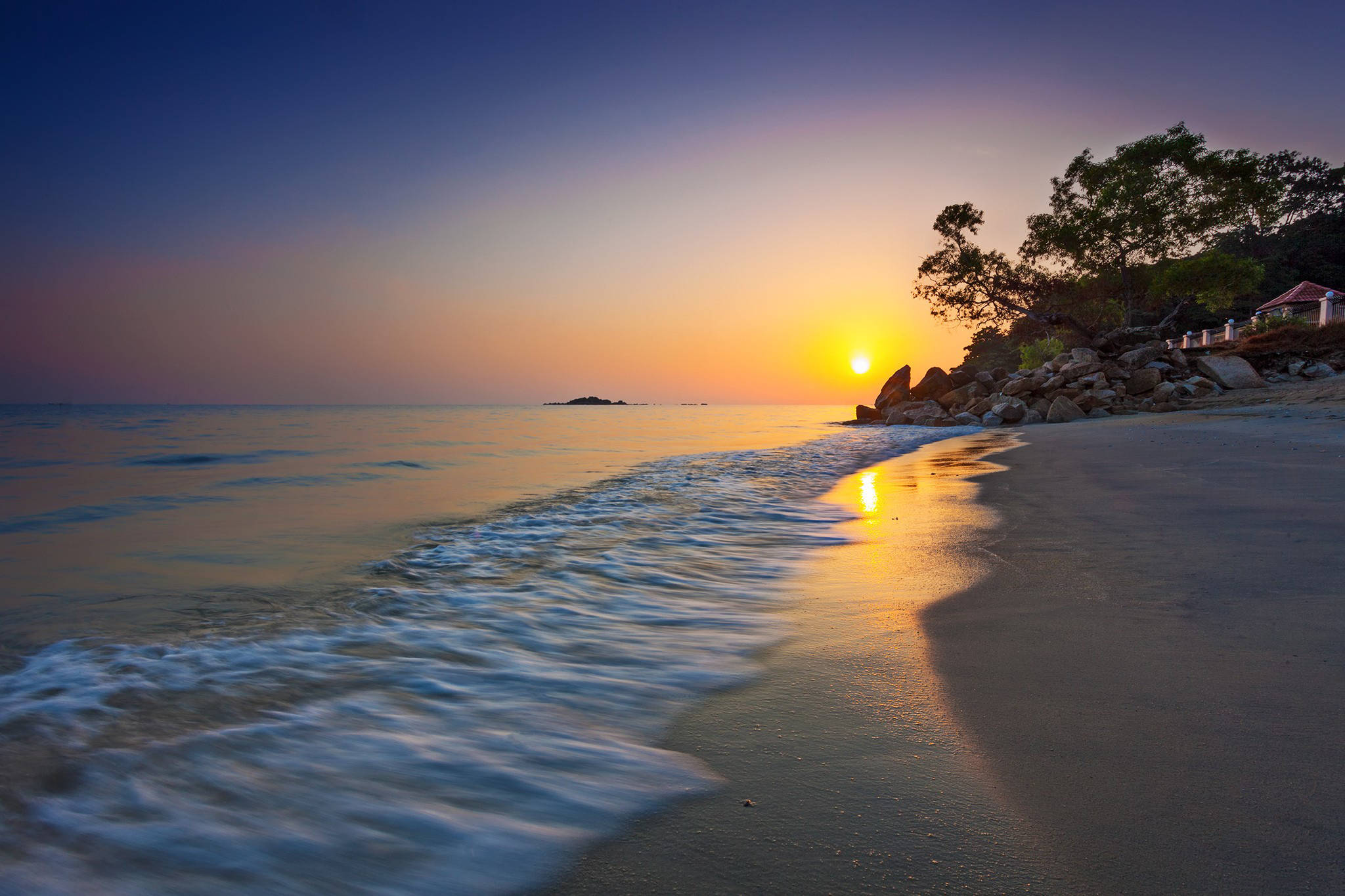 馬來西亞日落海灘 壁紙 — 金色夕陽照耀著平靜的海浪沖刷到沙灘上，沿岸有樹木和岩石。