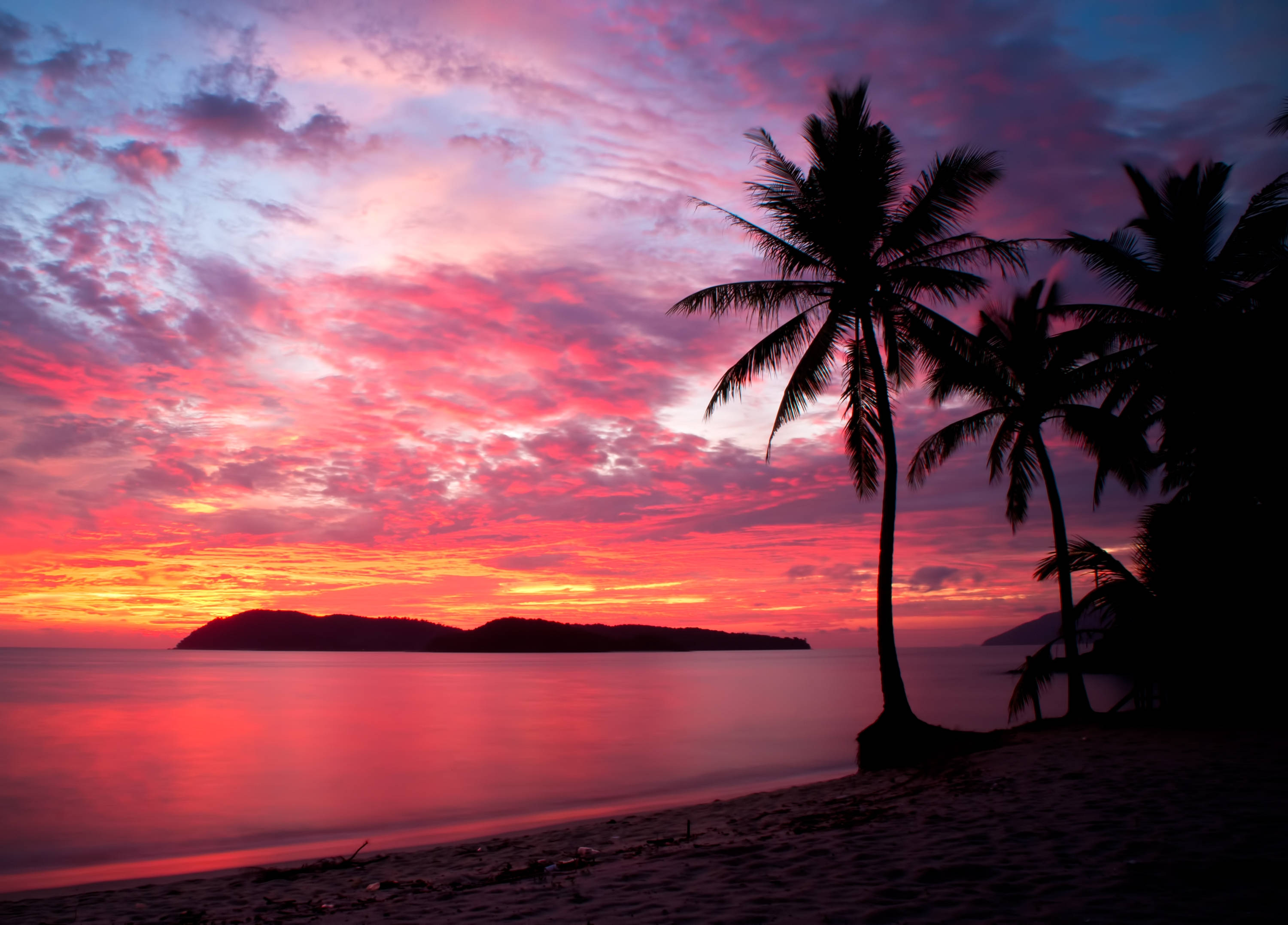 馬來西亞熱帶海灘粉紅夕陽 壁紙 — 剪影的棕櫚樹框住了平靜海面上遠方島嶼襯托下充滿活力的粉紅色和橙色日落。