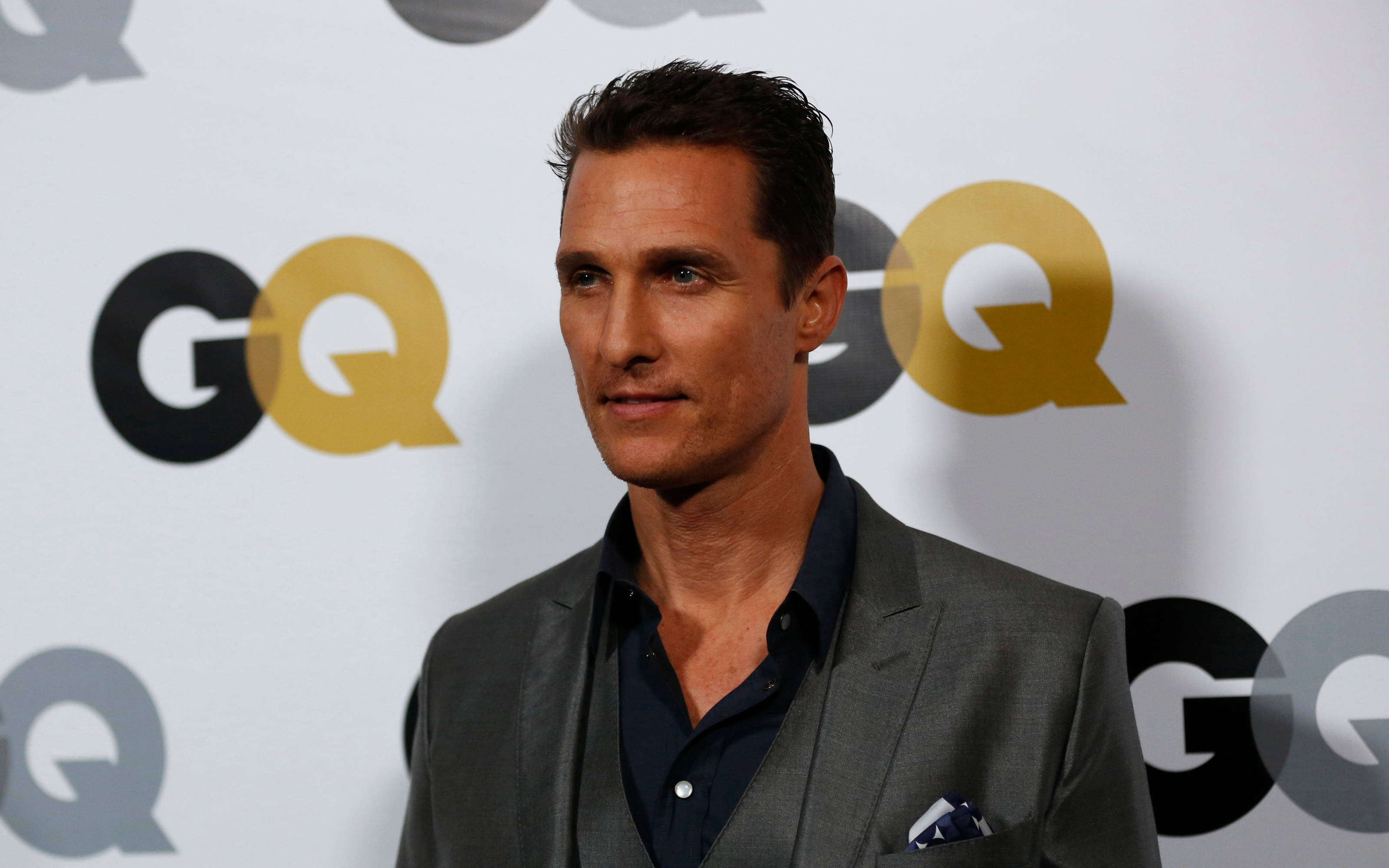馬修·麥康納為 GQ 雜誌拍攝 壁紙 — 一名身穿灰色西裝和深色襯衫的男子在活動中自信地站在重複的GQ標誌背景前。