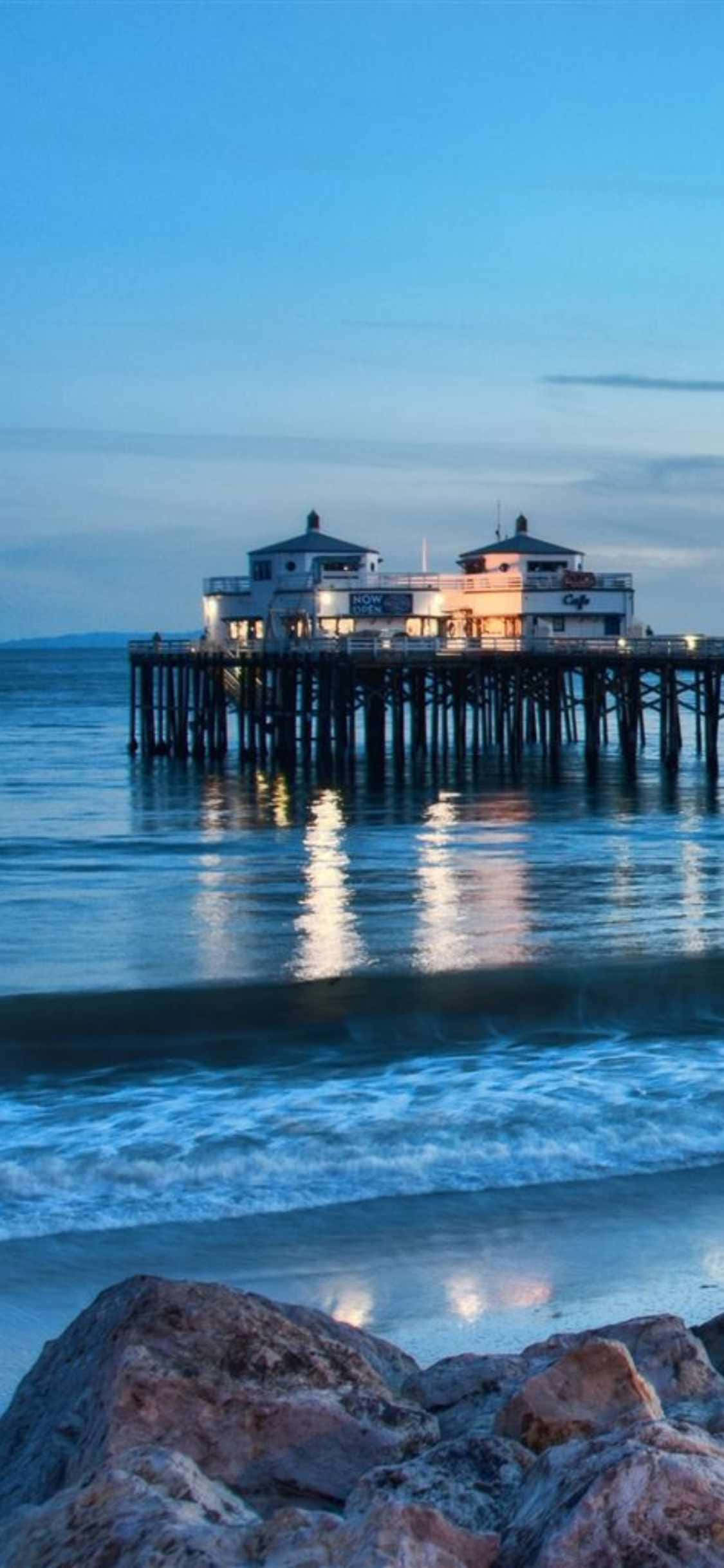 馬利布海灘日落美景 壁紙 — 一個寧靜的海岸碼頭，燈火通明的建築物倒映在平靜的藍色海水中，海浪在黃昏時拍打前景的岩石。