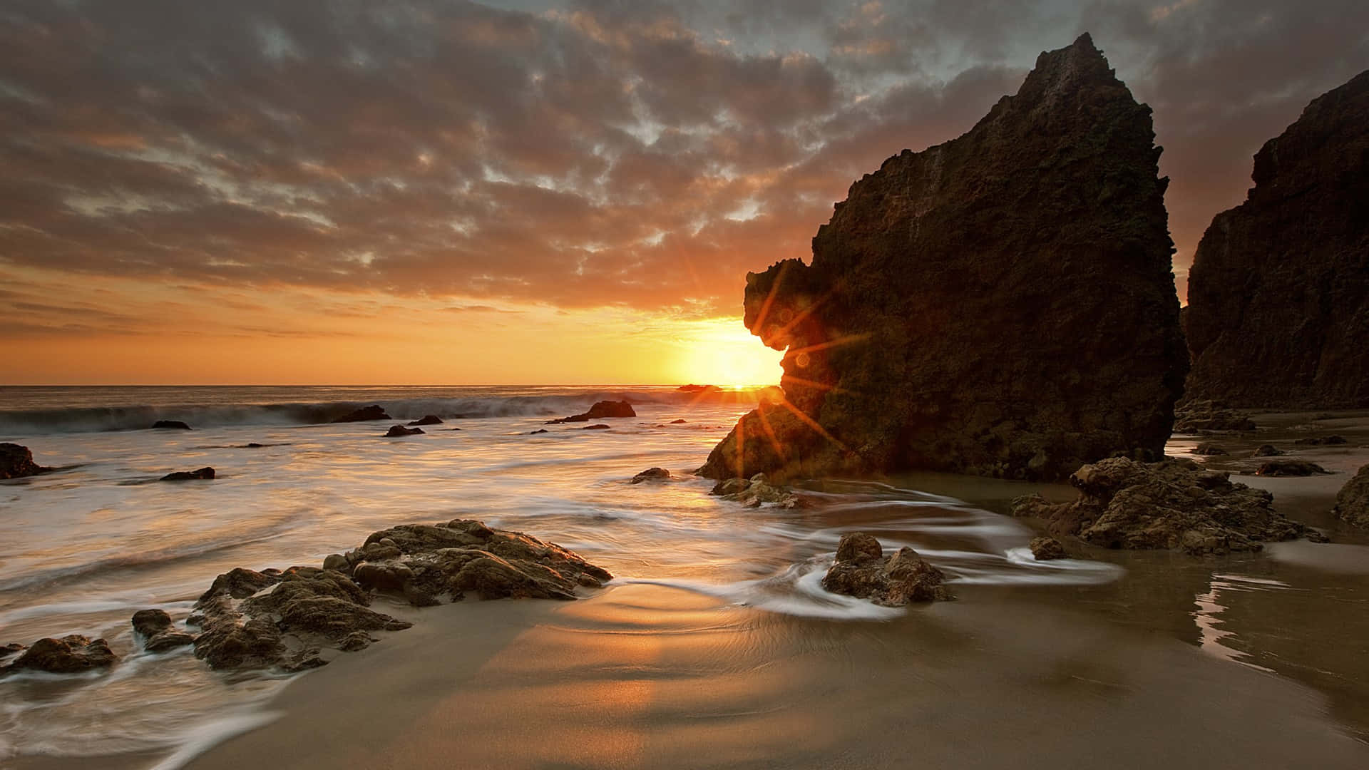 馬利布的岩石海灘 壁紙 — 夕陽透過岩石拱門閃耀，海浪在戲劇性的多雲天空下輕輕拍打海岸。