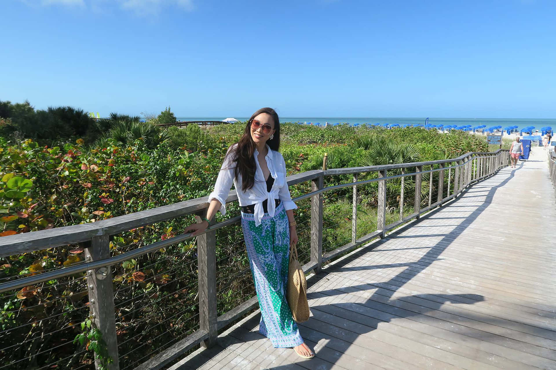 戴著太陽眼鏡、穿著圖案褲子的女士站在海灘附近的木製棧道上。