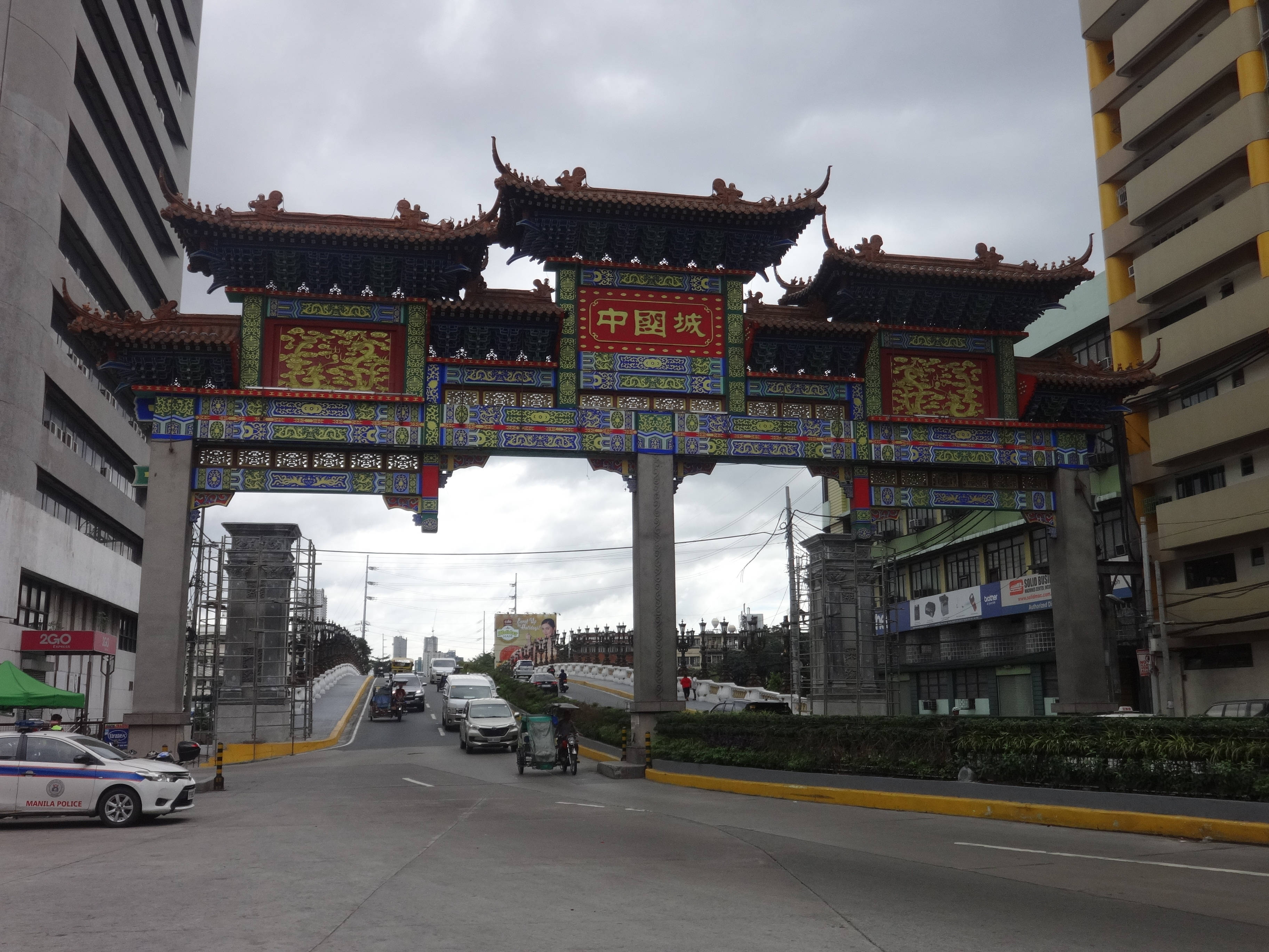 馬尼拉唐人街的活力脈搏 壁紙 — 一座宏偉華麗的中式牌坊，帶有紅色、金色和藍色的細節，矗立在繁忙的城市街道上，街道上有汽車和建築物，天空多雲。