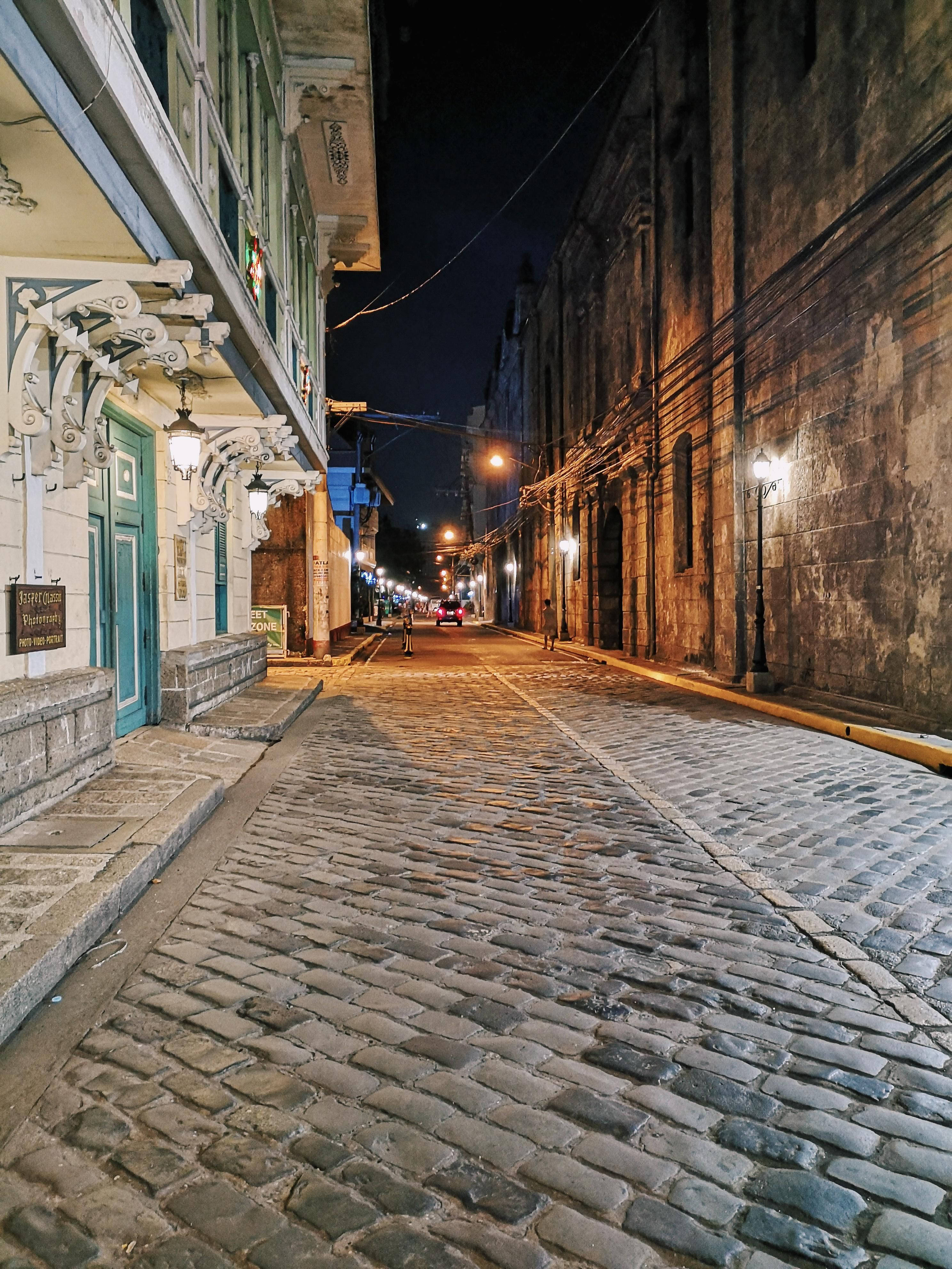 馬尼拉因特拉穆羅斯夜景燈光 壁紙 — 一條狹窄的鵝卵石街道，夜晚被溫暖的路燈和復古建築外牆照亮。