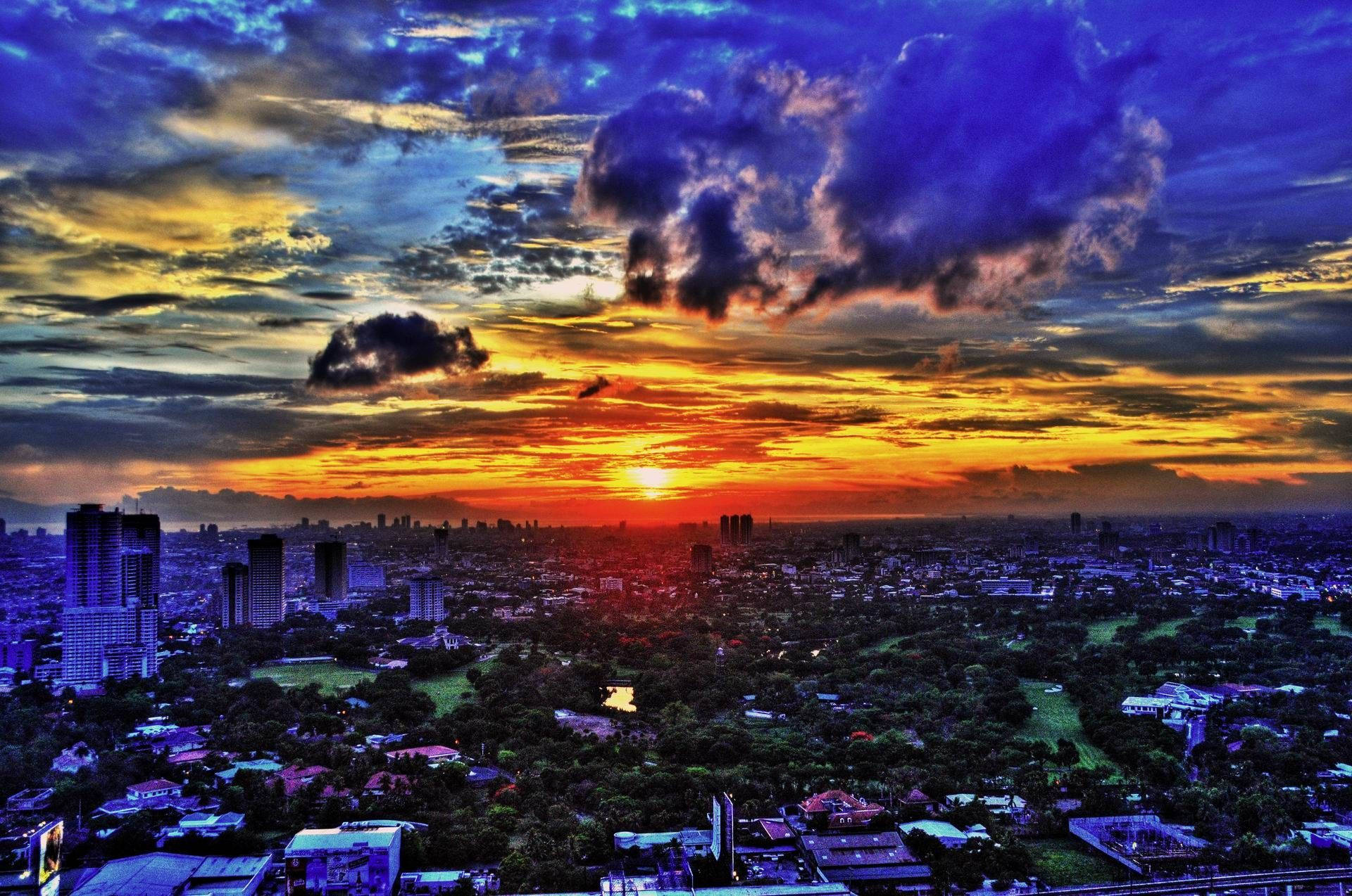 馬尼拉市日落 壁紙 — 夕陽西下，天空被染成火橙色和深紫色，下方是廣闊的城市景觀，高樓大廈與綠色公園交錯。