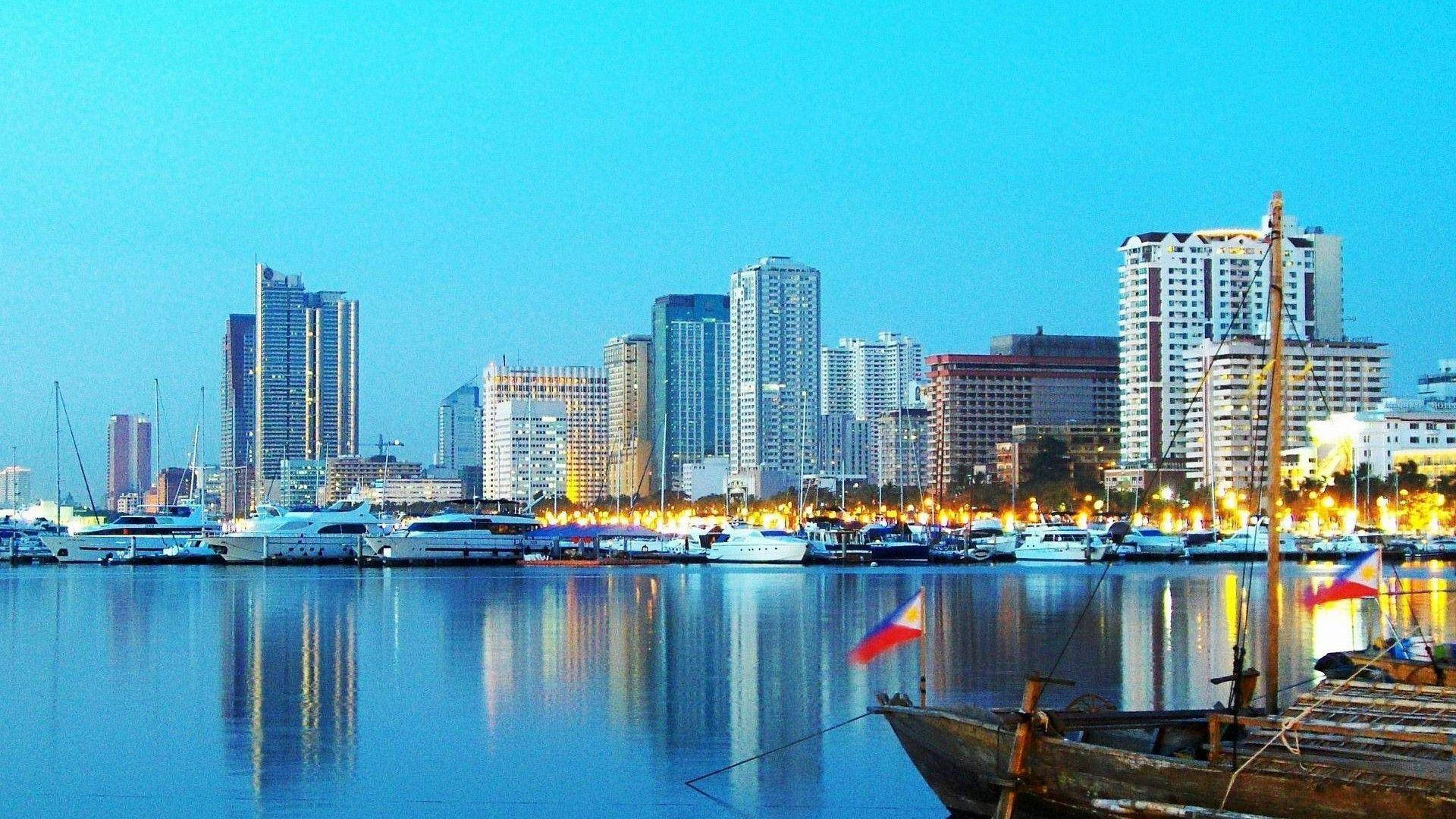 馬尼拉灣城市景觀 壁紙 — 寧靜的黃昏城市景觀，現代摩天大樓倒映在平靜的水面上，旁邊是傳統船隻。
