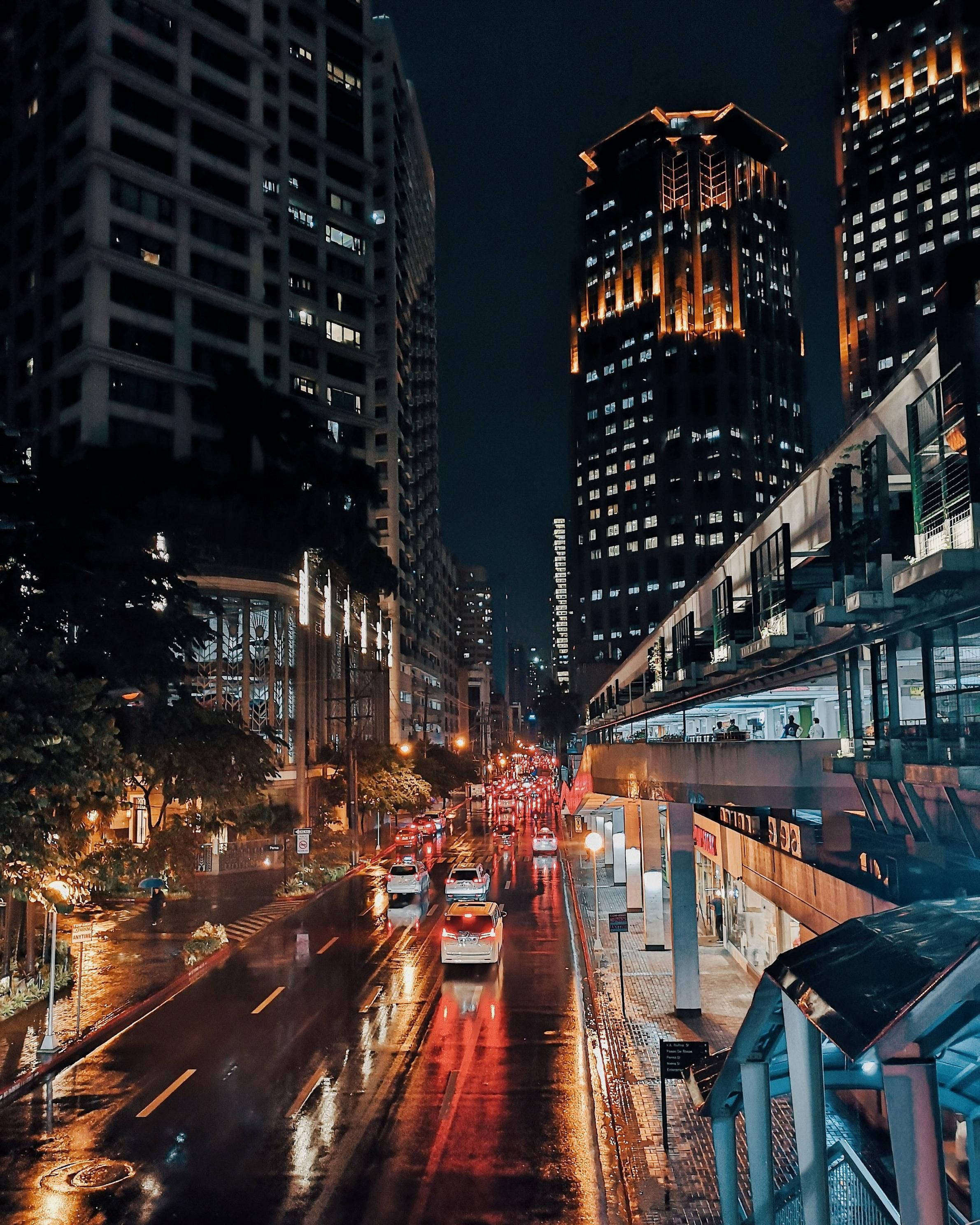 馬尼拉繁忙夜景燈光 壁紙 — 雨濕的夜晚城市街道，發光的汽車尾燈在濕漉漉的路面上反射，兩旁是高聳的照明摩天大樓。