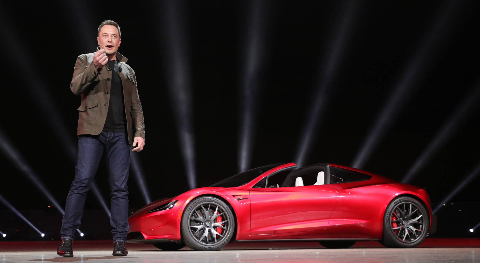 伊隆·馬斯克在燈光舞台上展示一輛紅色Tesla Roadster。 — 馬斯克 壁紙