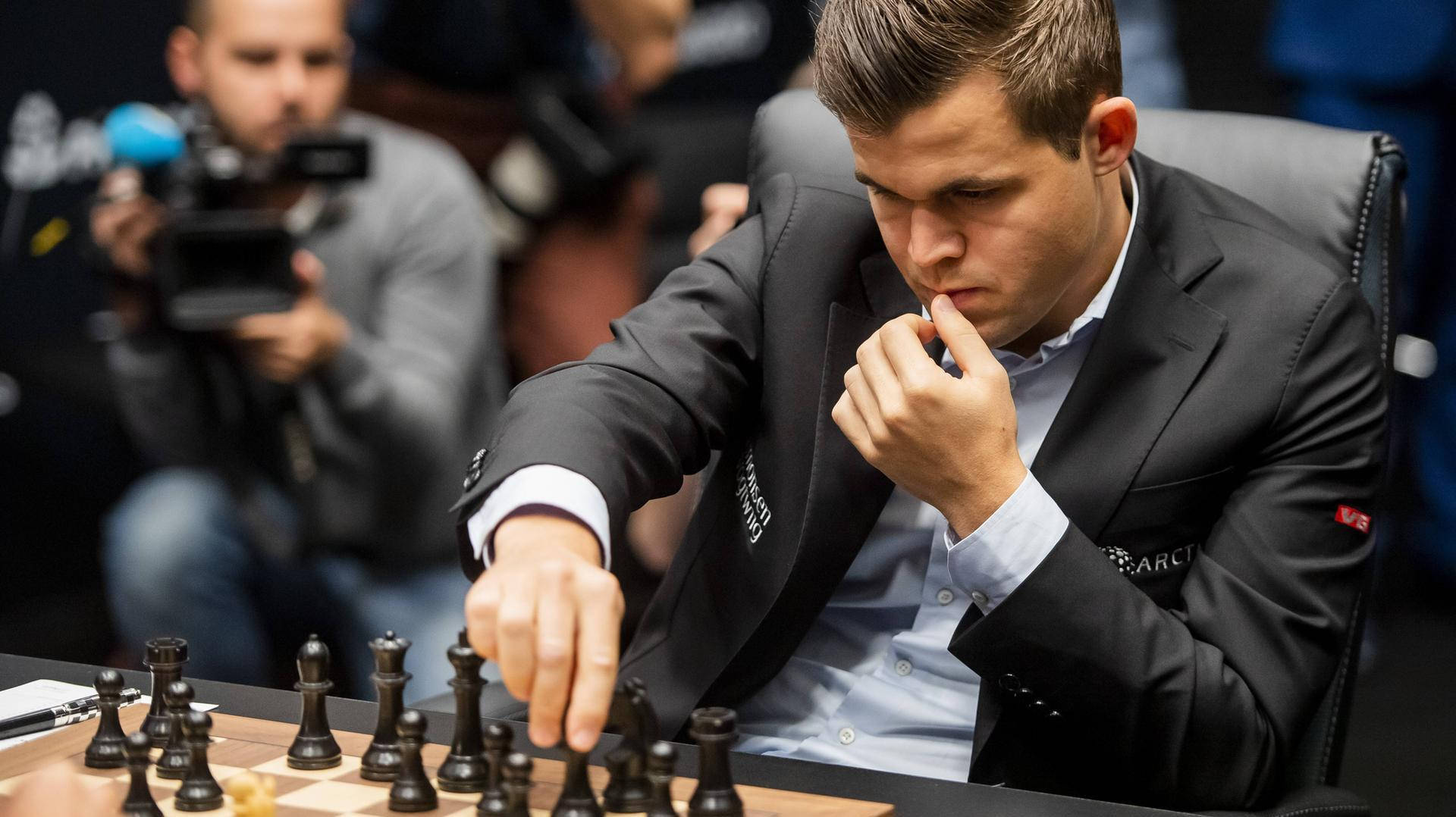 馬格努斯·卡爾森使用黑棋子比賽 壁紙 — 一位專注的西洋棋選手穿著黑色西裝，在木製棋盤上移動棋子，攝影師捕捉這緊張的時刻。