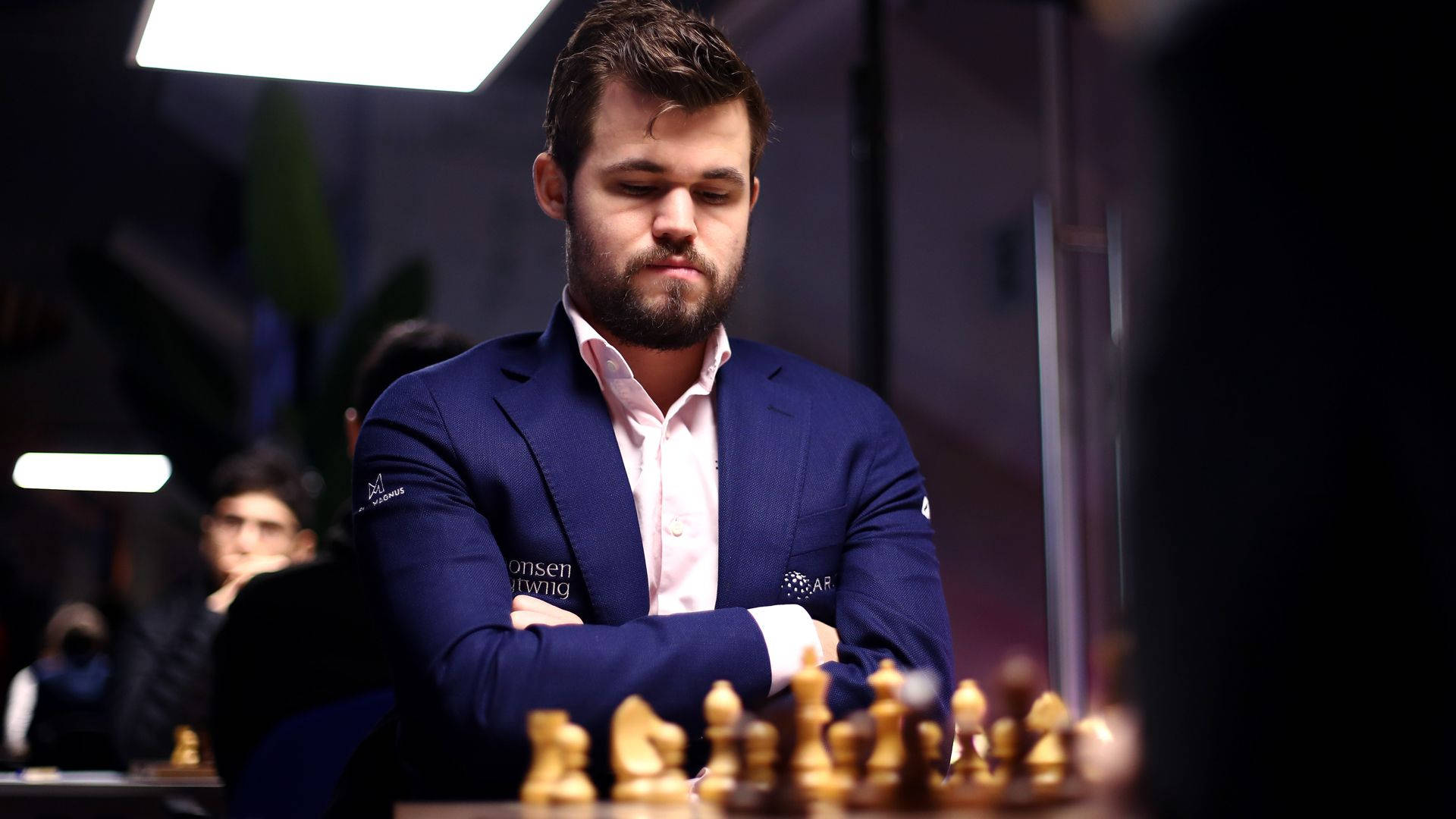 馬格努斯·卡爾森專注姿勢 壁紙 — 一位身穿藍色西裝、專注的棋手在比賽中思考下一步，背景有模糊的觀眾。