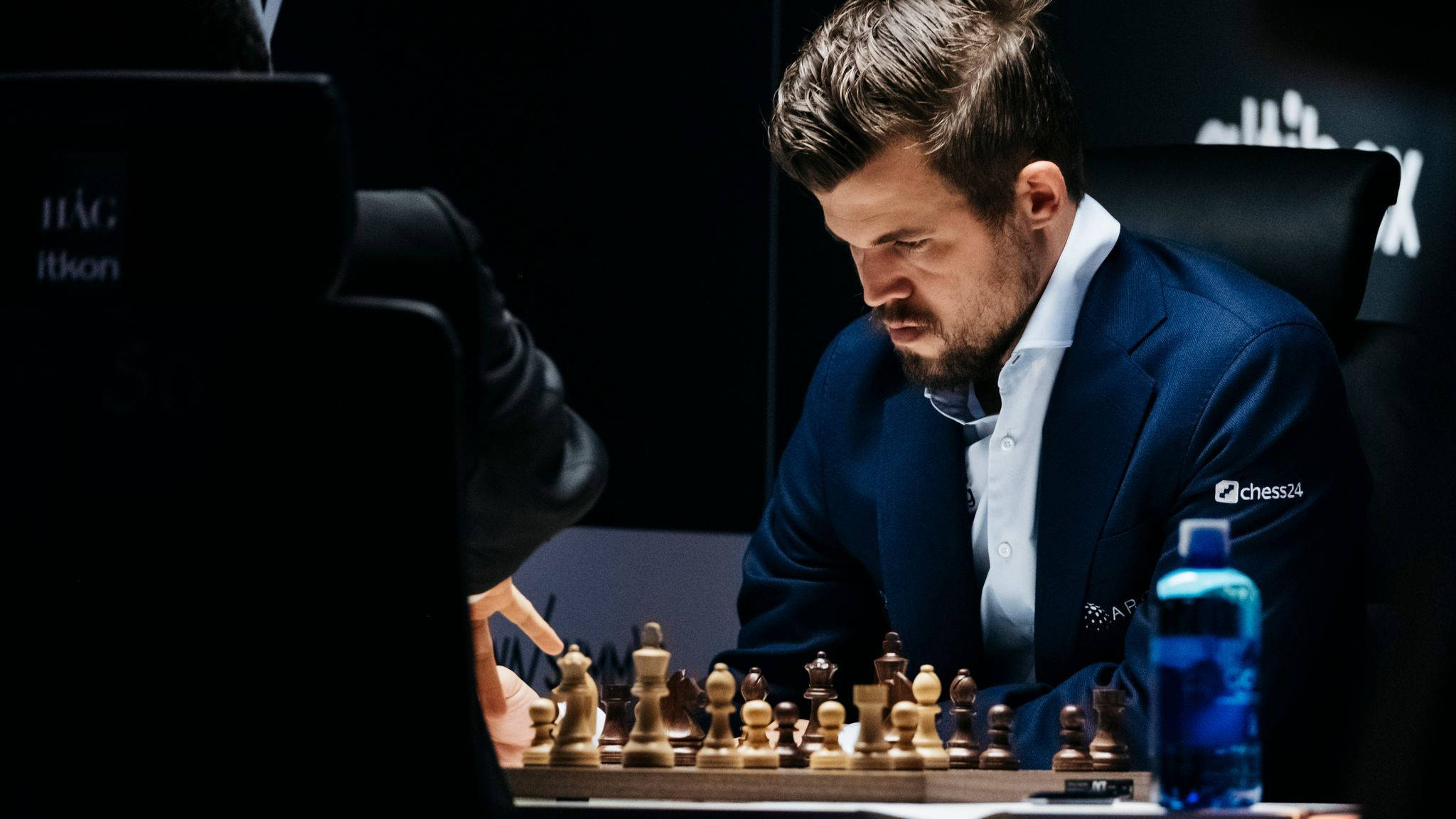 馬格努斯·卡爾森於棋局中專注 壁紙 — 一位身穿深色西裝、專注的國際象棋選手在昏暗的比賽環境中俯身於棋盤之上。
