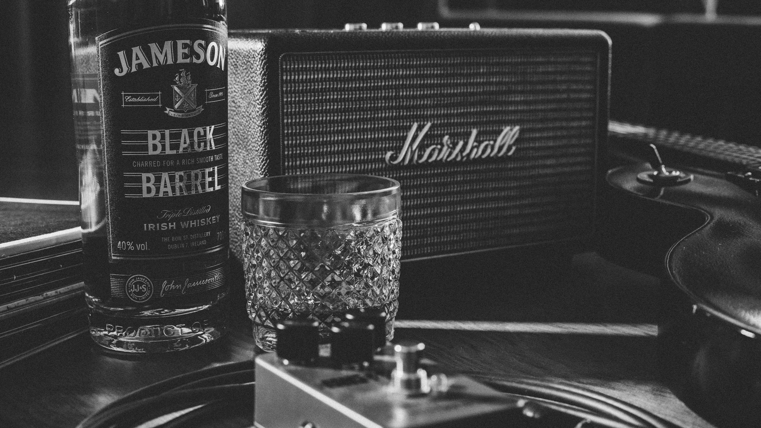 馬歇爾音箱搭配詹姆森威士忌 壁紙 — 木質表面上嘅 Jameson Black Barrel 酒瓶、酒杯同 Marshall 擴音器嘅單色靜物。