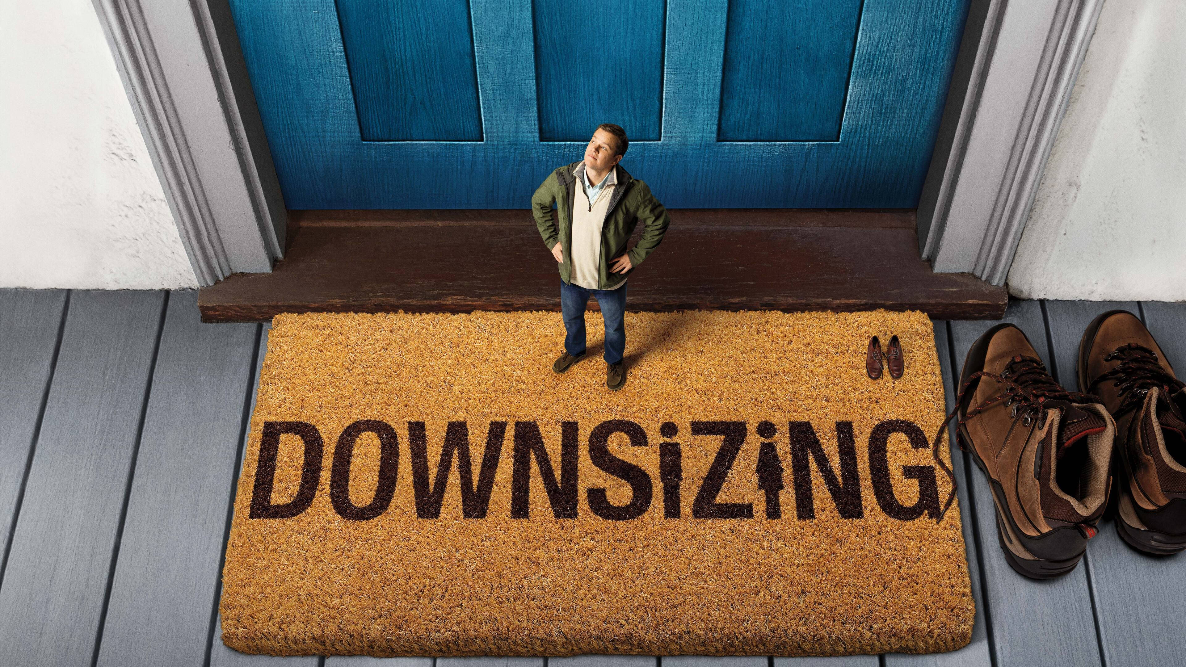 馬特·達蒙在《縮小人生》電影中 壁紙 — 一個男人站在寫著「DOWNSIZING」的門墊上，旁邊有一扇藍色門，鞋子整齊地放在一旁。