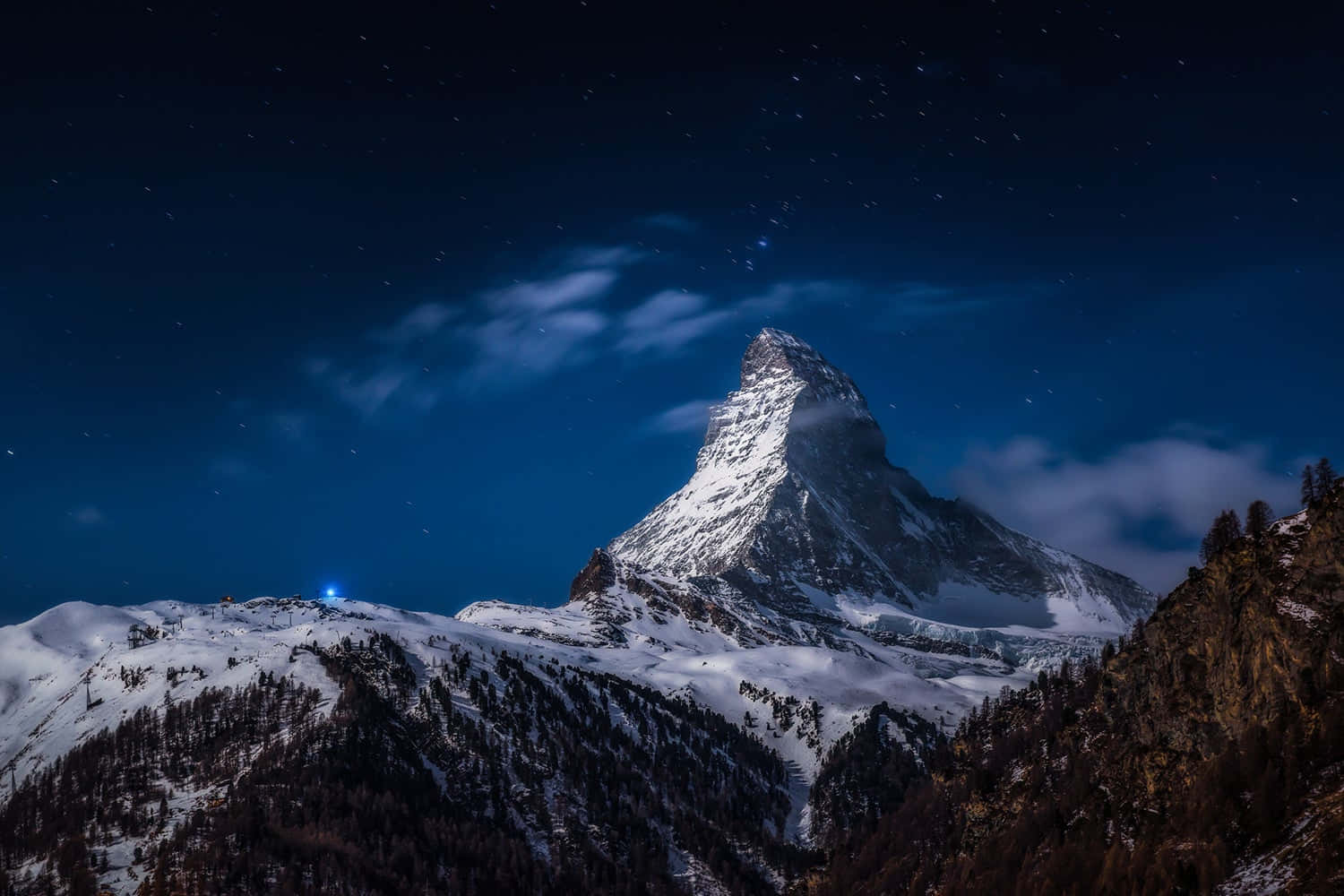 繁星點點的夜空下，白雪覆蓋的馬特洪峰，飄動的雲朵和下方的一盞藍光。