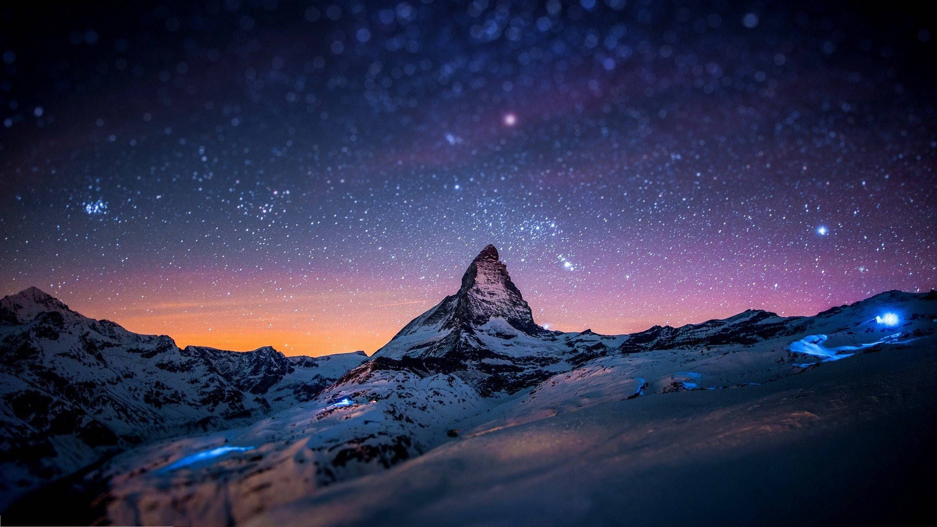 馬特洪峰的美麗夜景 壁紙 — 白雪覆蓋的馬特洪峰在星空暮色下升起，帶有紫橙色漸變和下方發光的藍色燈光。