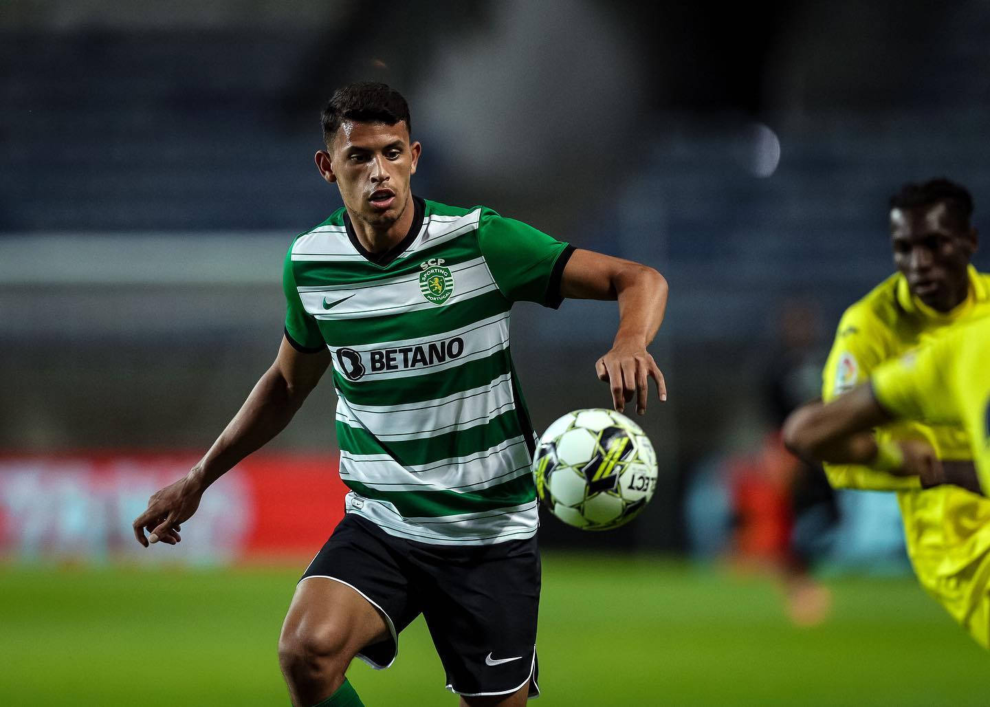 馬特烏斯·努內斯專注盯球 壁紙 — 一名身穿綠色和白色條紋的足球員正在盤球，而一名黃色球衣的後衛在球場燈光下的草地上逼近。