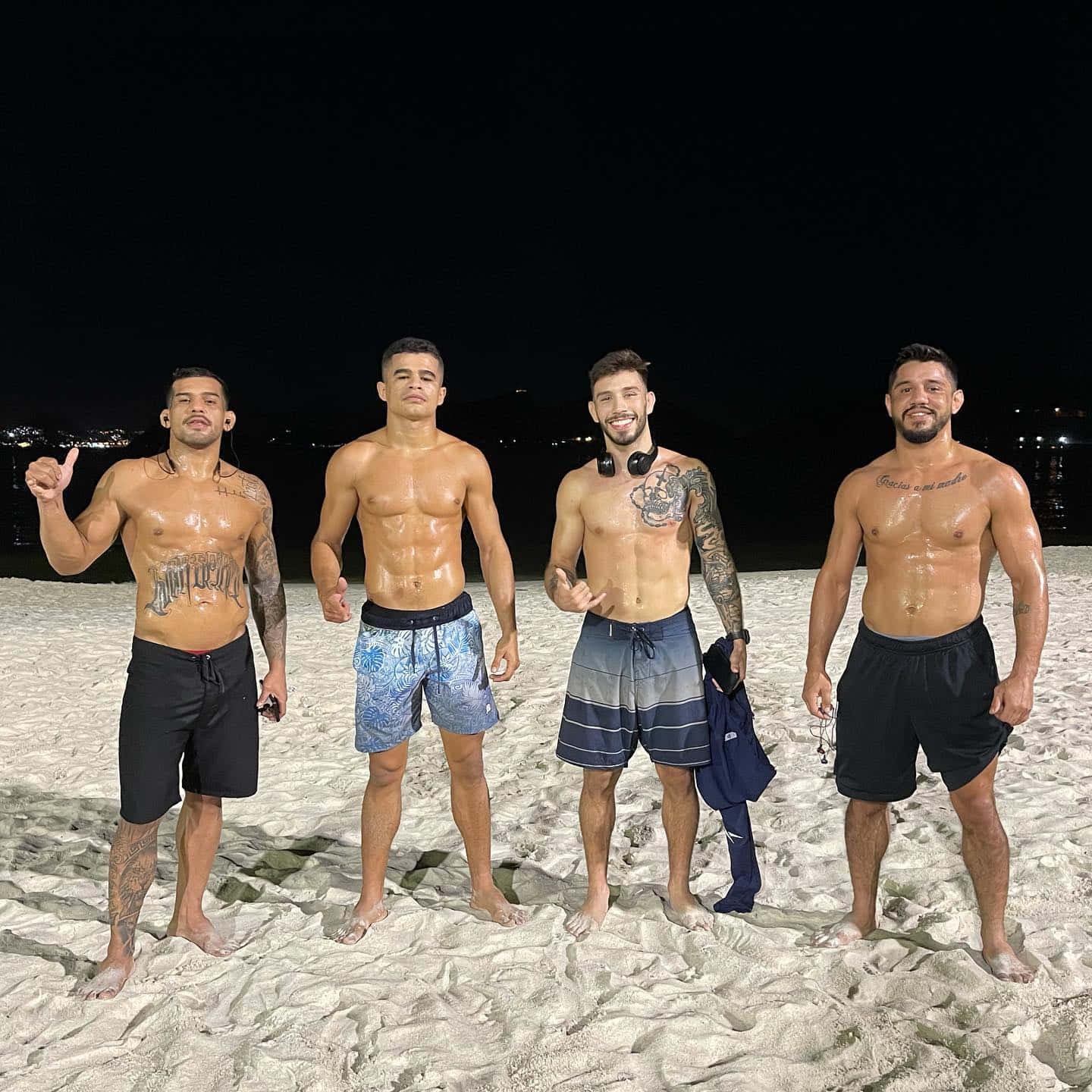 四個有紋身的赤膊男子在夜晚的海灘上以城市燈光為背景擺姿勢。