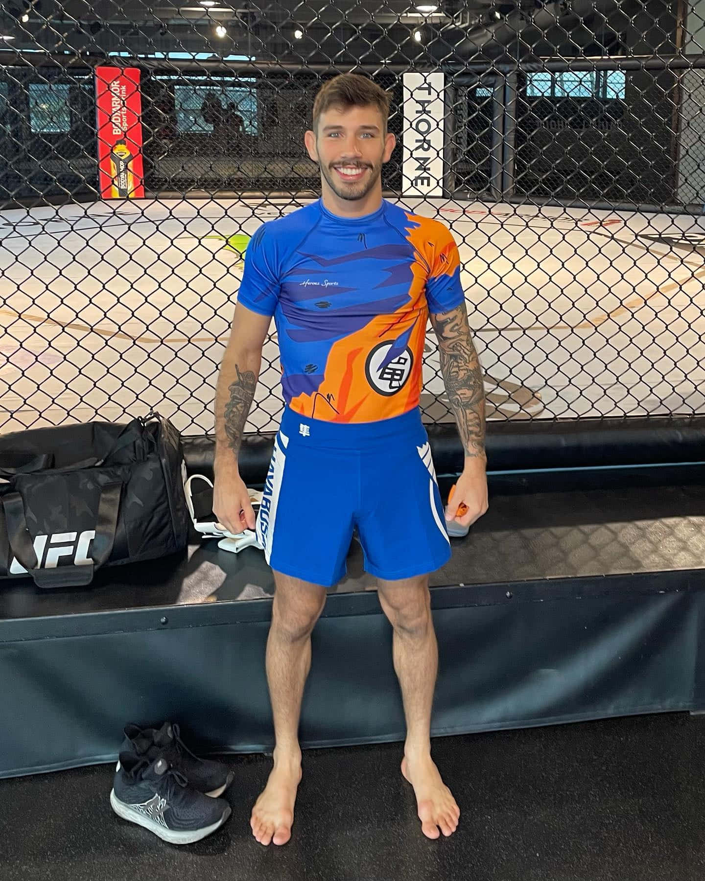 一位微笑的MMA戰士身穿藍色和橙色裝備，赤腳站在籠內。