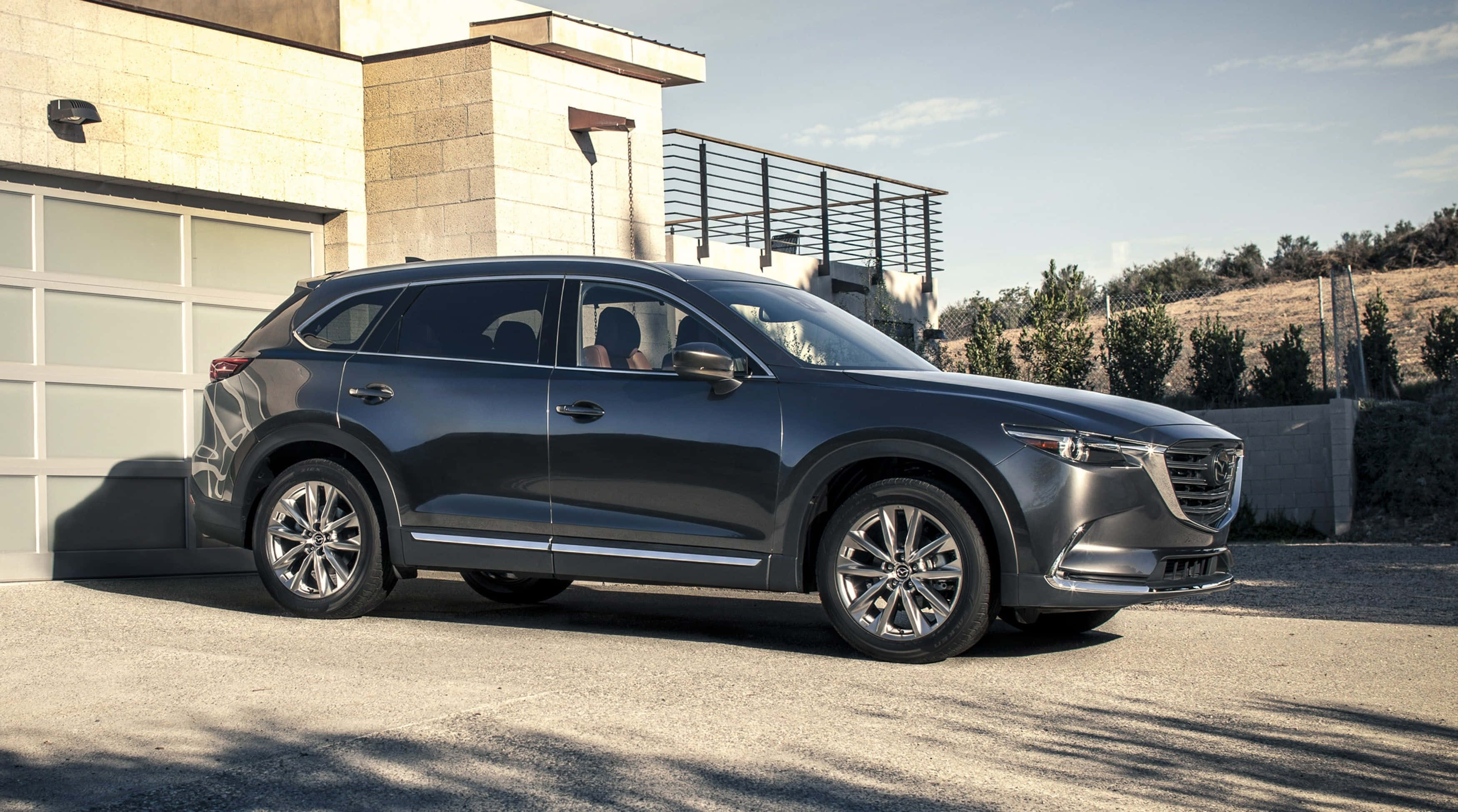 馬自達CX-9：極致駕駛體驗的視覺呈現 壁紙 — 一輛時尚的深灰色Mazda CX-9 SUV在明亮的日光下停在一棟現代石屋前。