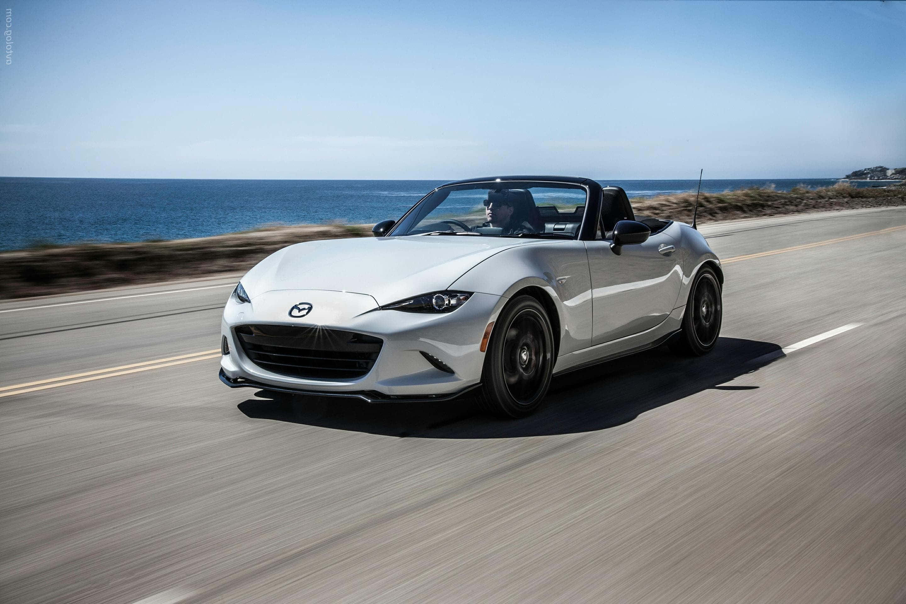 馬自達MX-5 Miata：速度與風格的完美結合 壁紙 — 一架時尚的銀色開篷跑車沿著海岸公路飛馳，海洋在晴朗的藍天下延伸至地平線。