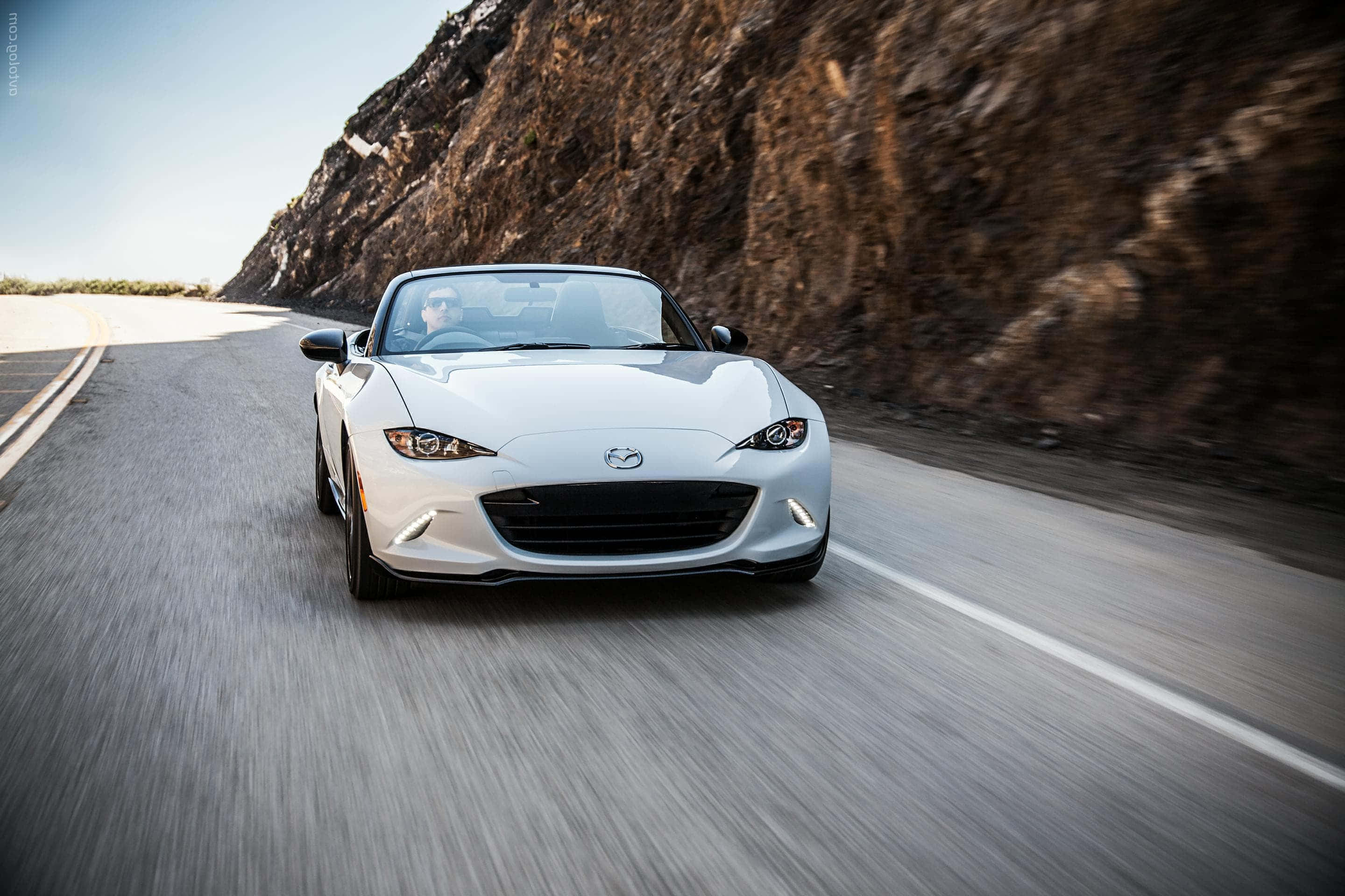 馬自達MX-5 Miata在開放道路上展現優雅風采 壁紙 — 一輛銀色敞篷跑車沿著蜿蜒的山路疾馳，帶有模糊的動態和岩石峭壁。