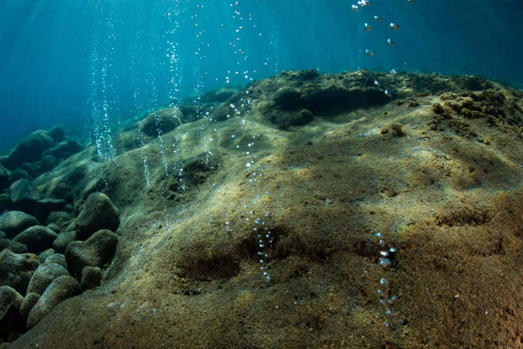 馬里亞納海溝的壯麗風景 壁紙 — 陽光穿過碧綠色的海水，照亮了岩石露頭附近的沙質海床和上升的氣泡。