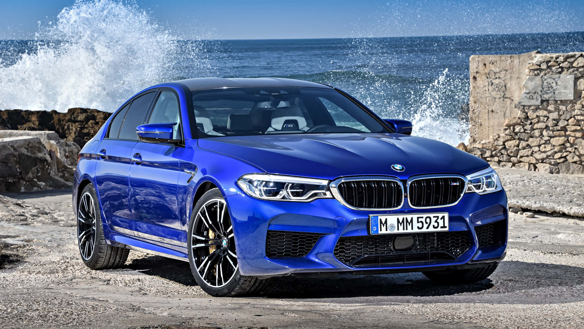 藍色BMW M5房車停泊在岩石海岸附近，海浪拍打。