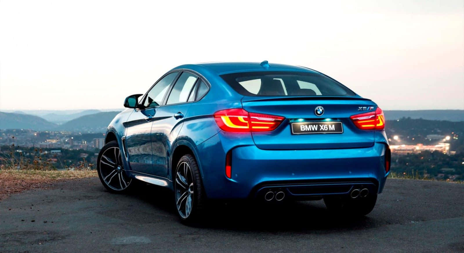駕馭 BMW X6 M，征服開闊道路 壁紙 — 一輛鮮藍色BMW X6 M轎跑車停在俯瞰城市景觀的山頂上，在黃昏時分亮起尾燈。