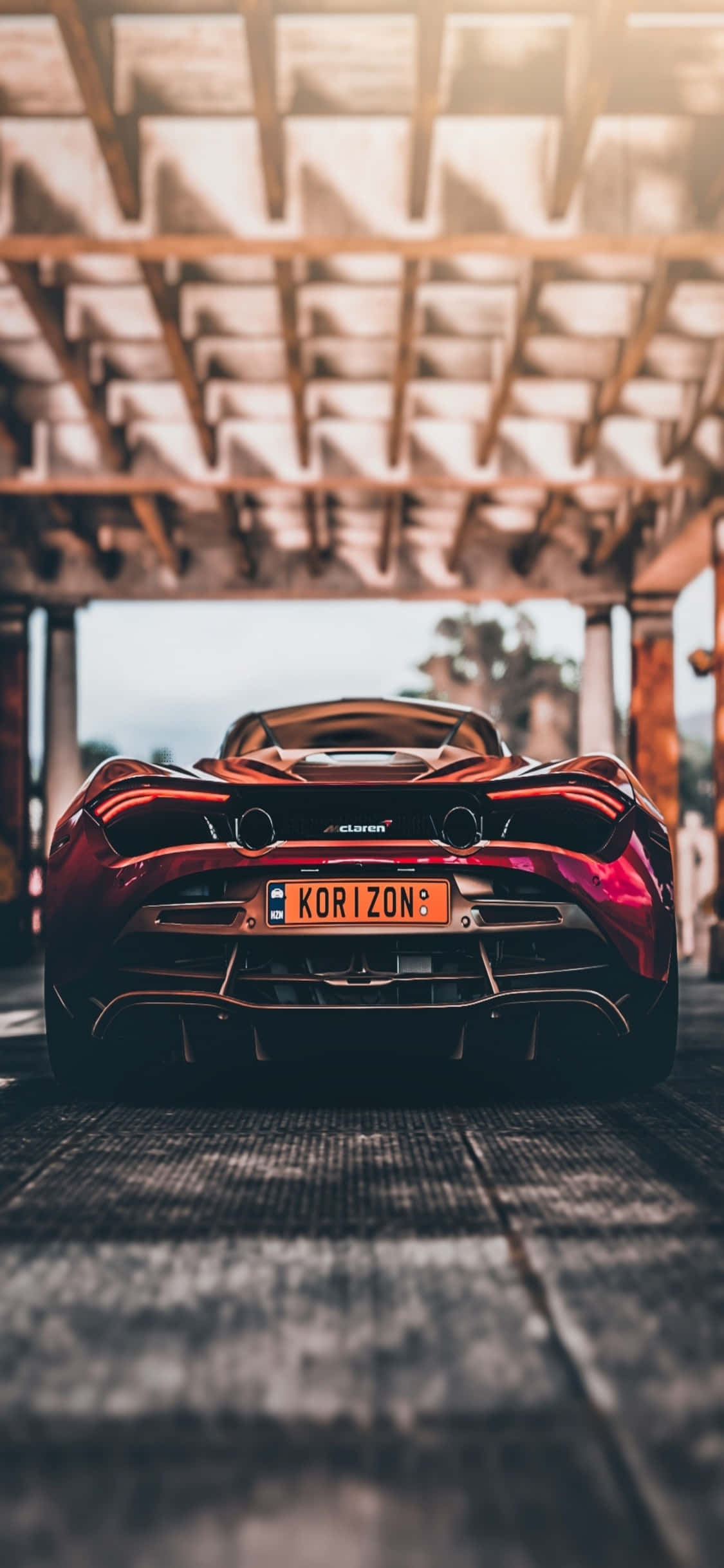 駕馭 iPhone Xs McLaren 720s，升級您的風格 壁紙 — 一輛光澤的紅色McLaren跑車停放在木結構下，陽光透過上方的橫樑照射進來。