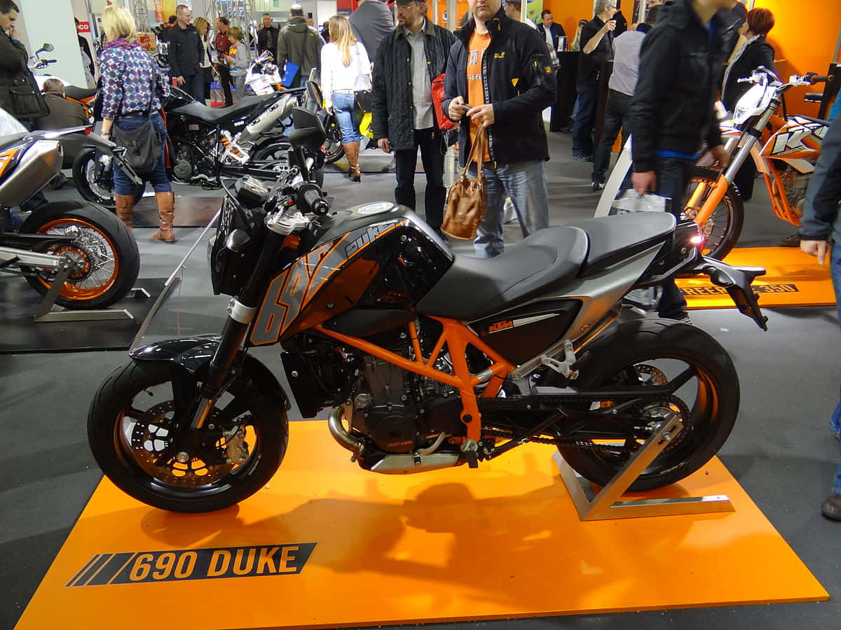 駕馭 KTM Duke 探索開放道路 壁紙 — 一輛時尚的橙色和黑色KTM 690 Duke摩托車在擁擠的室內展覽中展示，與會者走動觀看。