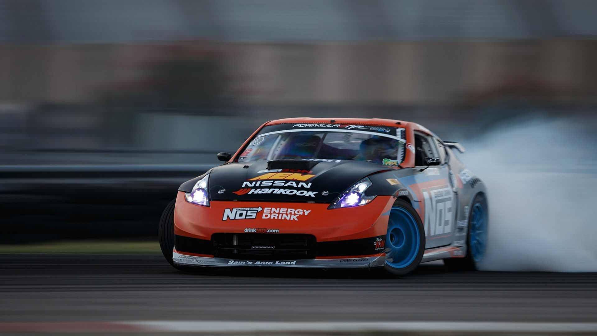 駕馭 Nissan 350Z，震撼道路 壁紙 — 一輛充滿活力的橙色和黑色漂移車，配備藍色輪胎，在賽道彎角處激進地冒煙。