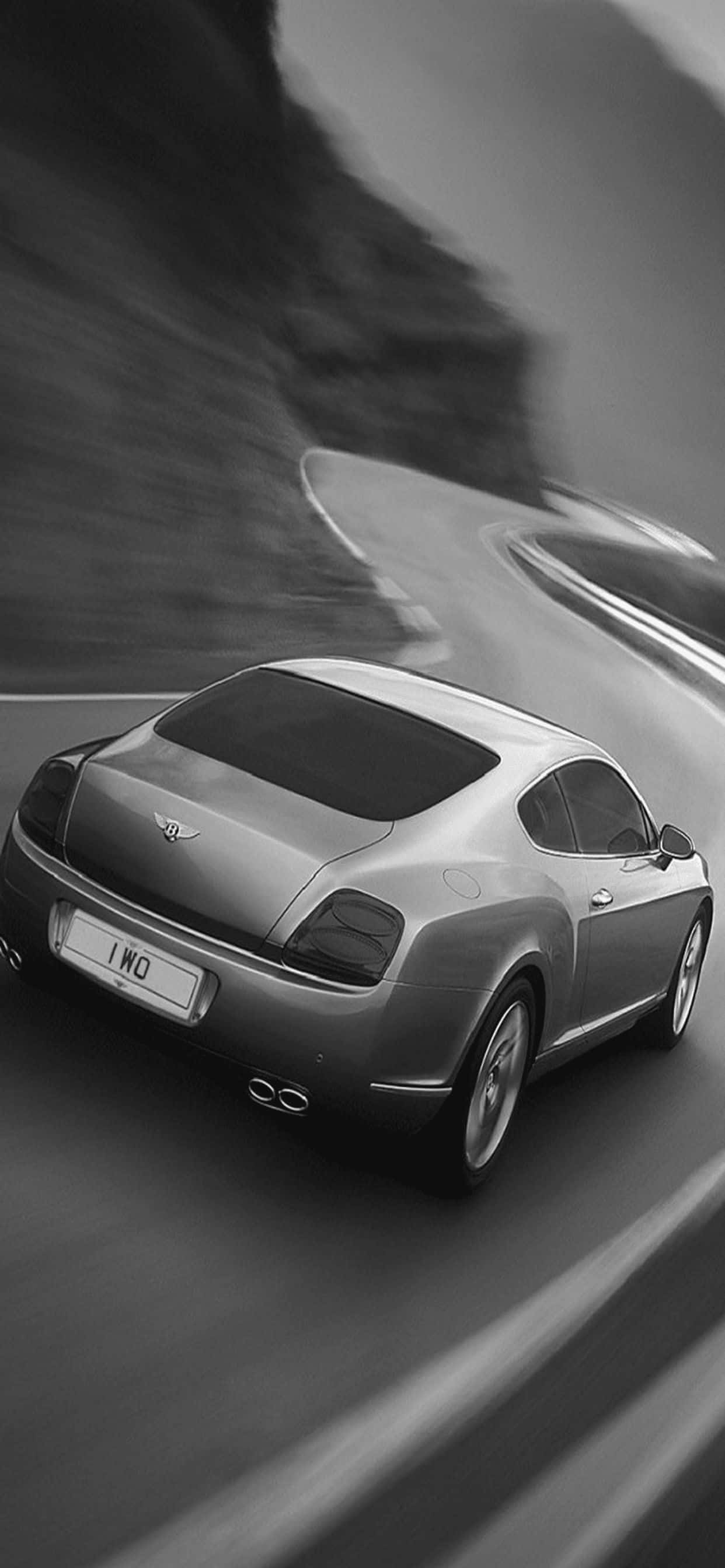 駕馭奢華：體驗 iPhone XS Max Bentley 壁紙 — 一輛時尚的銀色賓利在蜿蜒的山路上疾馳，以戲劇性的黑白動態模糊效果捕捉。