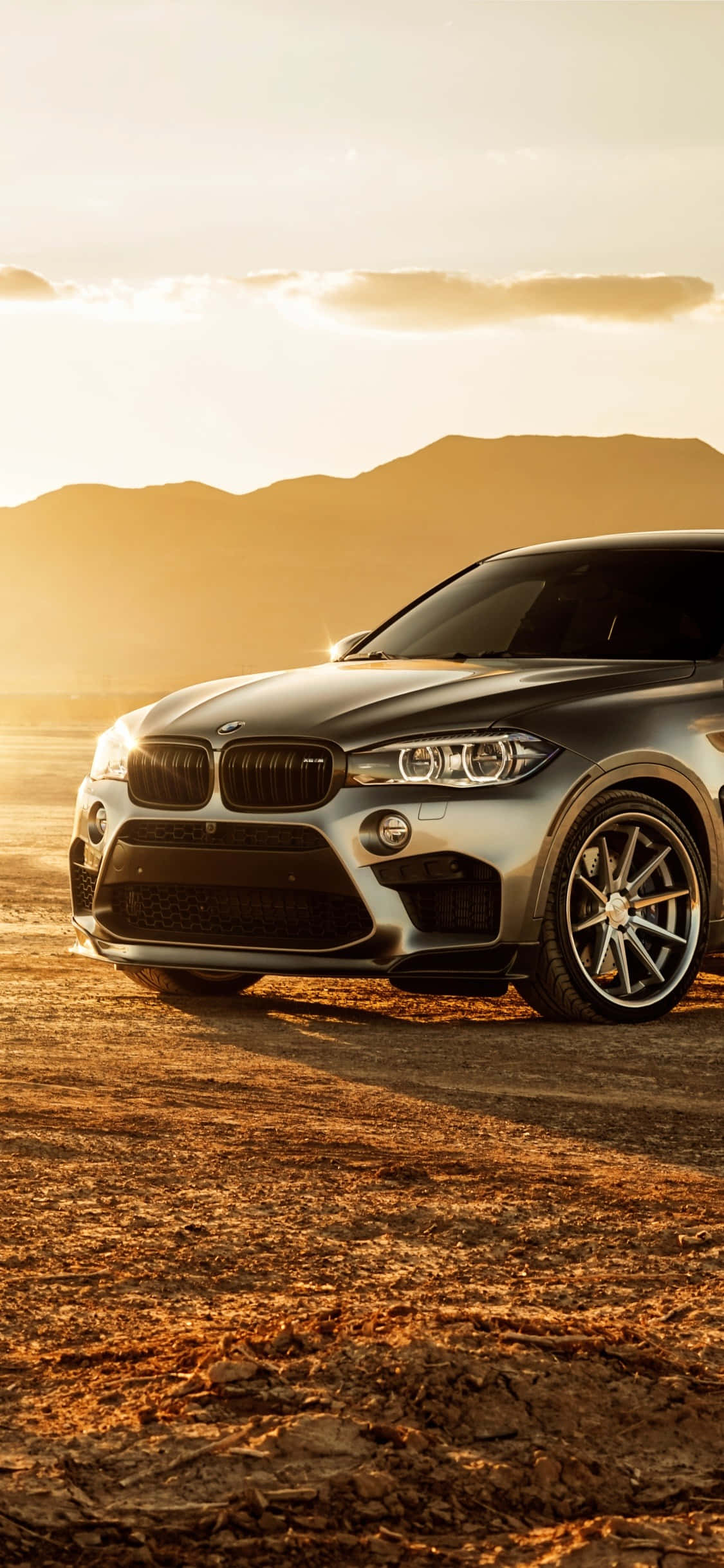 駕馭生活奢華 — iPhone X 與 BMW 壁紙 — 一架型格銀色BMW X6 SUV停泊在塵土飛揚的地形上，背景係金色夕陽同遠山。