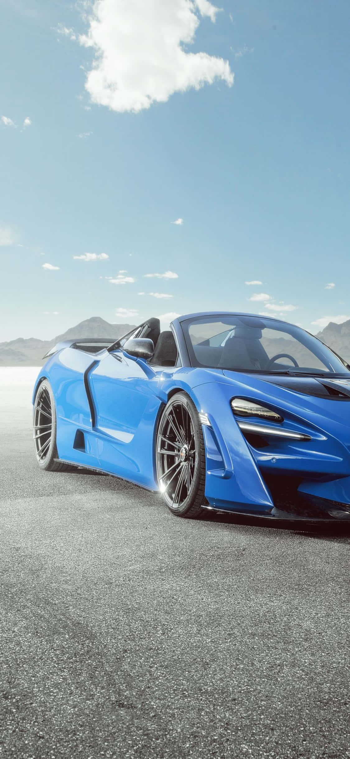 駕馭街頭風采，iPhone Xs 與 McLaren 720s 壁紙 — 一架充滿活力的藍色開篷跑車停泊在陽光照耀的瀝青路上，遠山在晴朗的天空下。