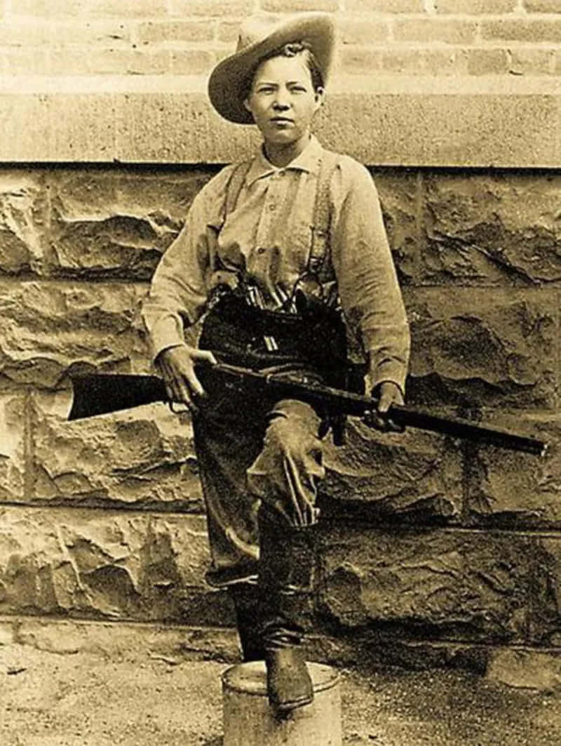 身穿歷史軍服的女性手持步槍坐在石牆前的木桶上。