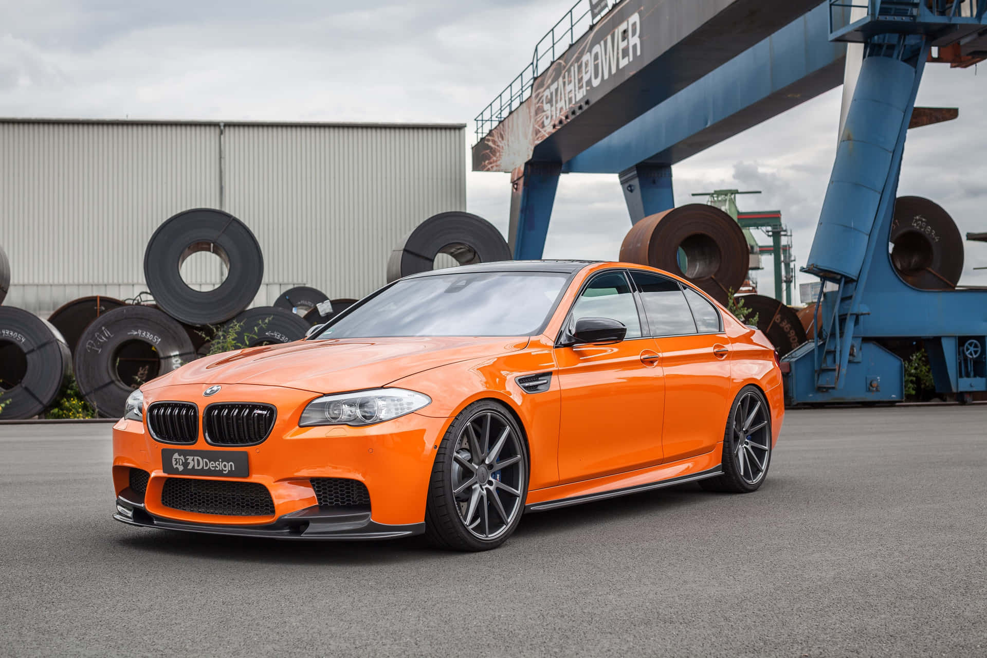 橙色BMW M5停放在工業場地，有鋼卷和藍色起重機。